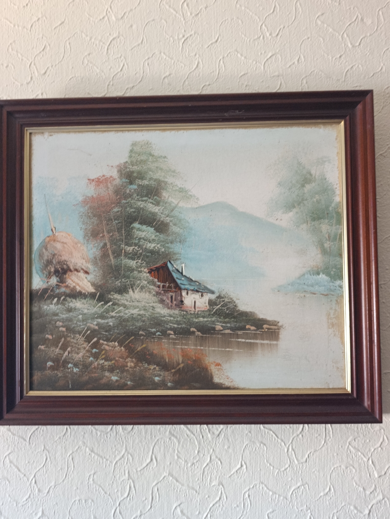 This is a photograph of a framed oil painting depicting a pastoral landscape. The painting features a small, rustic house with a blue roof situated on the bank of a body of water, possibly a lake or river. To the left of the house, a large, conical haystack is visible. The background shows misty hills and trees, with a soft, hazy atmosphere. The painting is framed in a dark, wooden frame with a gold inner border. The artwork is hung on a textured white wall. No specific location in Setúbal, Portugal, can be determined from the painting itself.