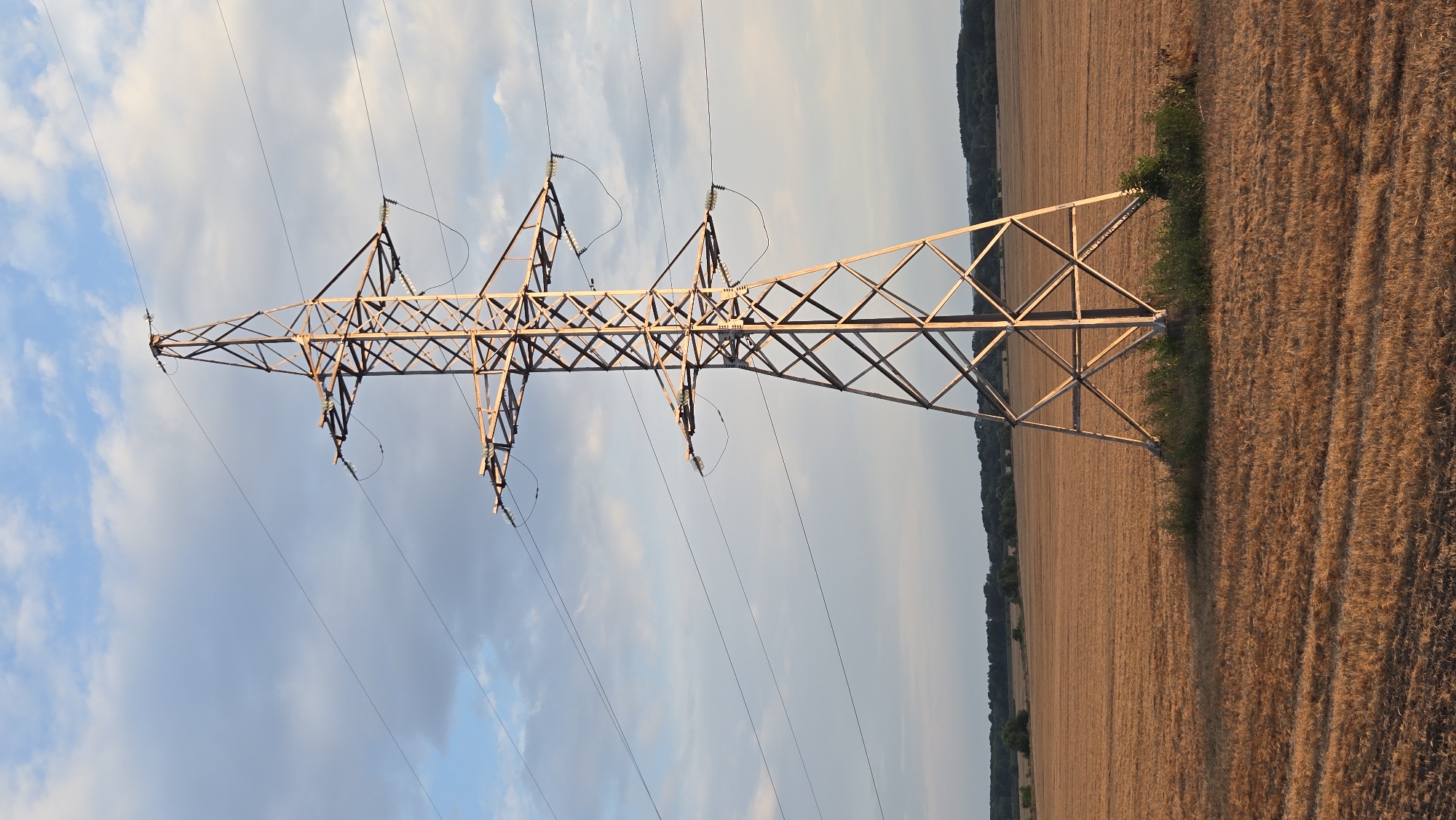 A tall, lattice-style electricity pylon stands in the center of a harvested wheat field under a sky with scattered clouds. The pylon is made of metal and has several crossbeams where electrical wires are suspended. The field is a uniform golden-brown color, indicating it has recently been mowed or harvested. In the distance, a line of trees forms the horizon, with a subtle indication of a hilly landscape beyond. The lighting suggests it is either early morning or late afternoon, with a warm glow illuminating the scene. The image appears to have been taken in a rural setting, possibly near Suklieia, Moldova, given the context provided. There are no people or animals visible in the frame.