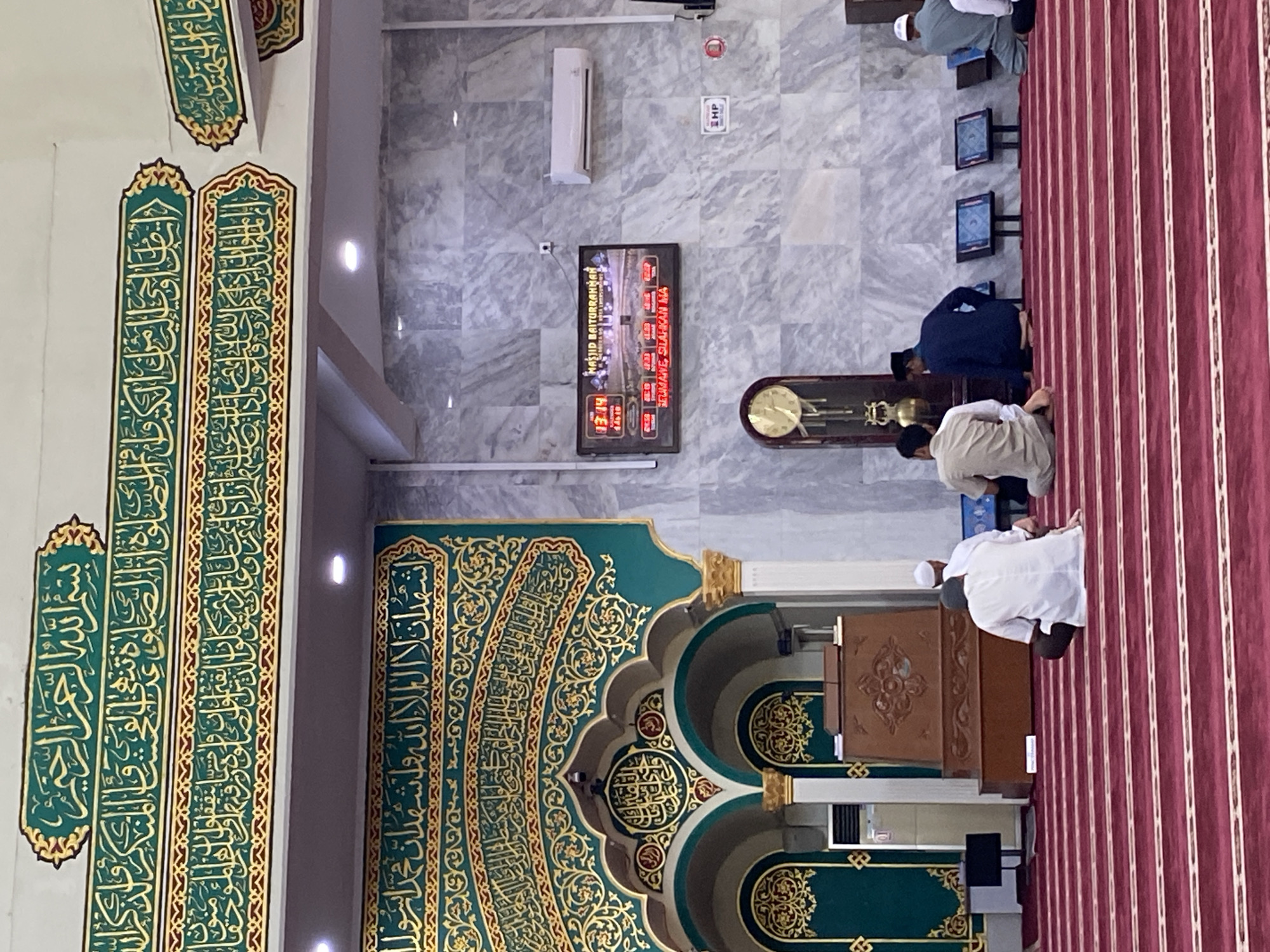 The image depicts the interior of the Baiturrahman Grand Mosque in Lhokseumawe, Indonesia. Several men are seated on a red carpeted floor in prayer. In the background, a digital display shows the time and mosque name, "MASJID BAITURRAHMAN". Arabic calligraphy adorns the walls and archways. A large grandfather clock stands to the left of the digital display. The walls are covered in marble-patterned material. An air conditioning unit is mounted on the wall above the digital display. The scene captures a moment of quiet devotion within the mosque.