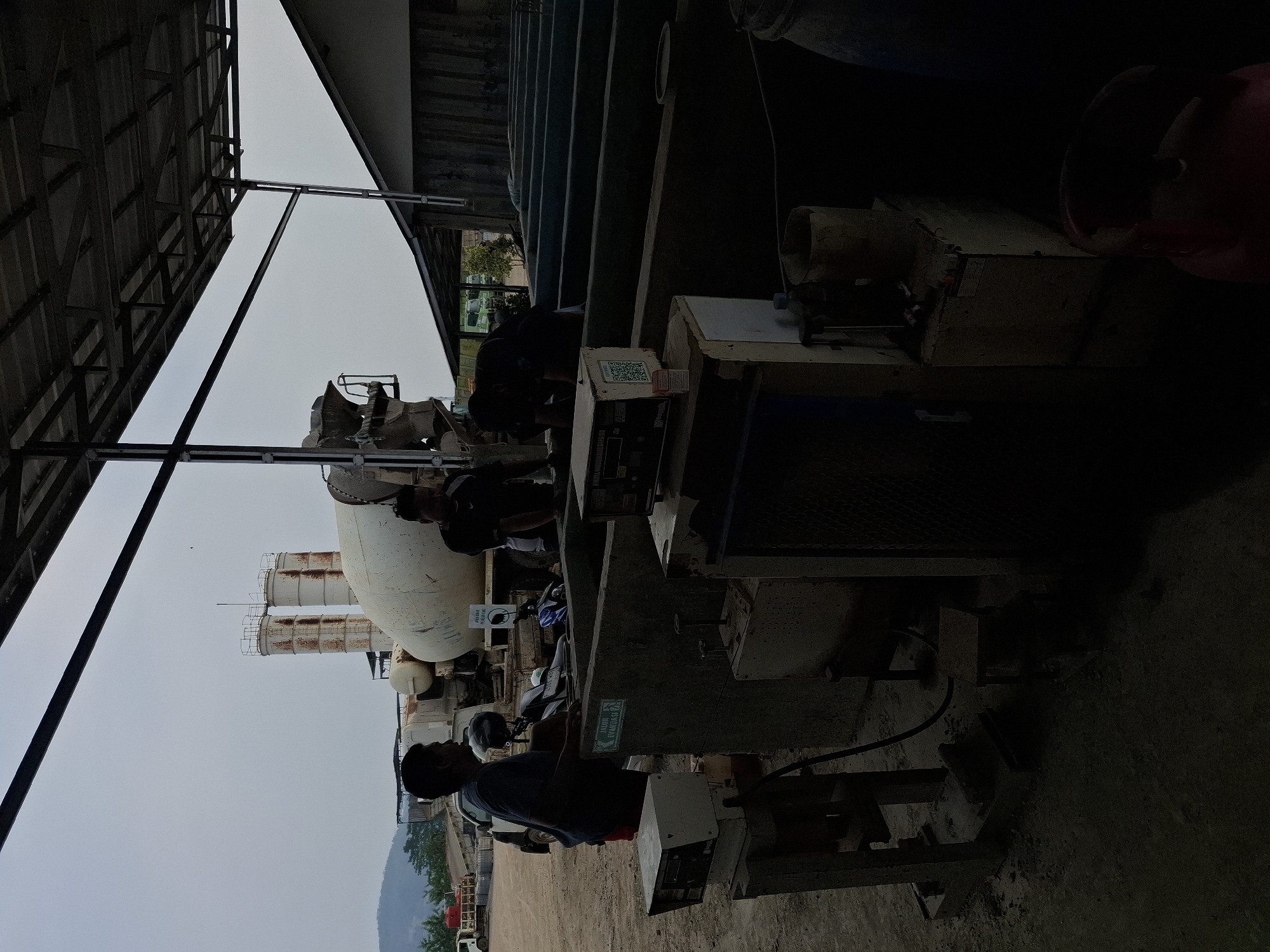In Kutasirna, Indonesia, a concrete mixing plant operates under an open-sided metal roof. Two men are present. One man, wearing a dark blue t-shirt, stands near a concrete trough, looking towards the left of the frame. Another man, in a black t-shirt, is partially visible behind him, also facing left. A cement mixer truck with a rotating drum is parked in the background, and two tall concrete silos are visible further beyond. In the foreground, several pieces of equipment are situated: a scale with a digital display, a concrete mixer, and various containers. The ground appears to be dirt and gravel.