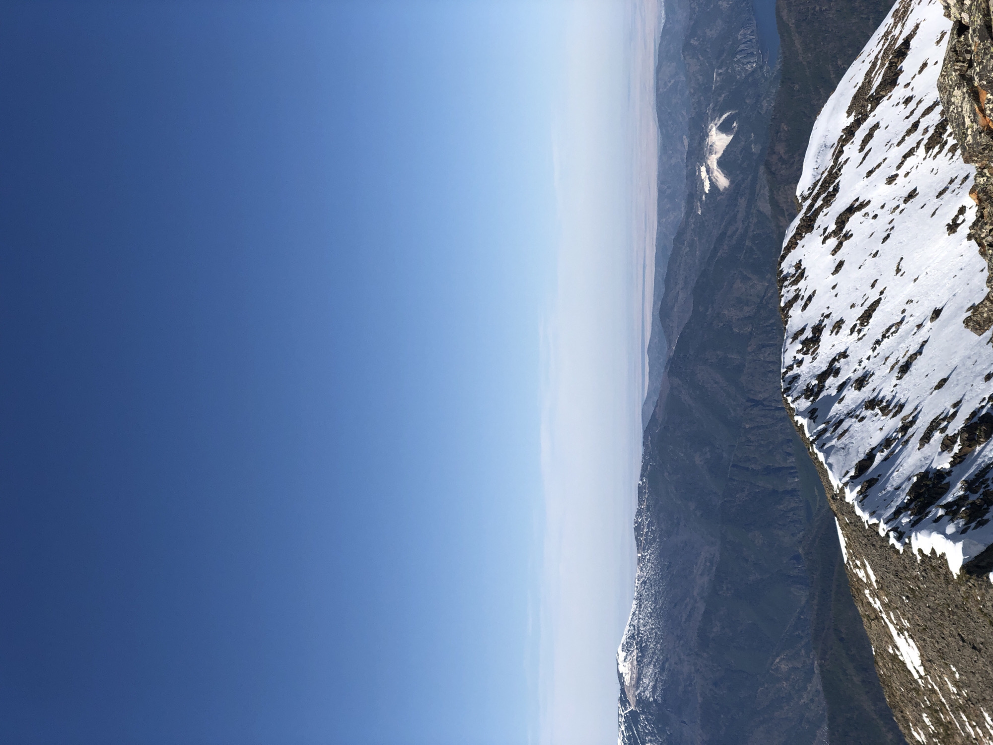 The image presents a mountain landscape with a clear blue sky occupying the majority of the frame. The right side of the image shows a steep mountain slope, partially covered with snow, indicating a high altitude or cold climate. The rocky terrain is visible through patches in the snow cover. In the distance, beyond the mountain range, there is a hazy horizon, suggesting a flat or low-lying area. Based on the prompt, the location is in Sayanogorsk, Russia, but specific details confirming this from the image alone are not available. No people or objects other than natural elements are discernible.
