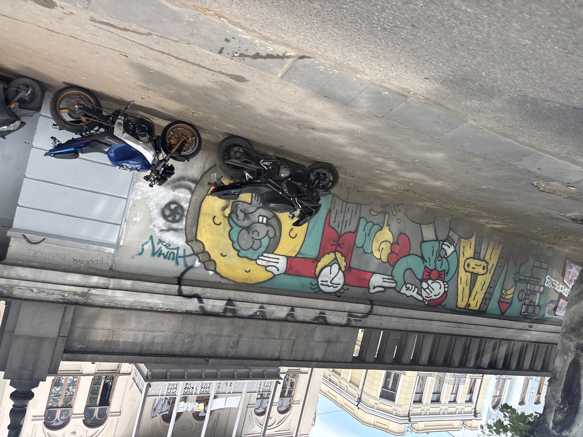 The image shows an urban scene in Tbilisi, Georgia. The view is upward toward a bridge or overpass support structure. Two motorcycles, one blue and one black, are parked on the edge of the structure. The support structure has a mural of cartoon figures in various colors. Behind the bridge, buildings with windows are visible.