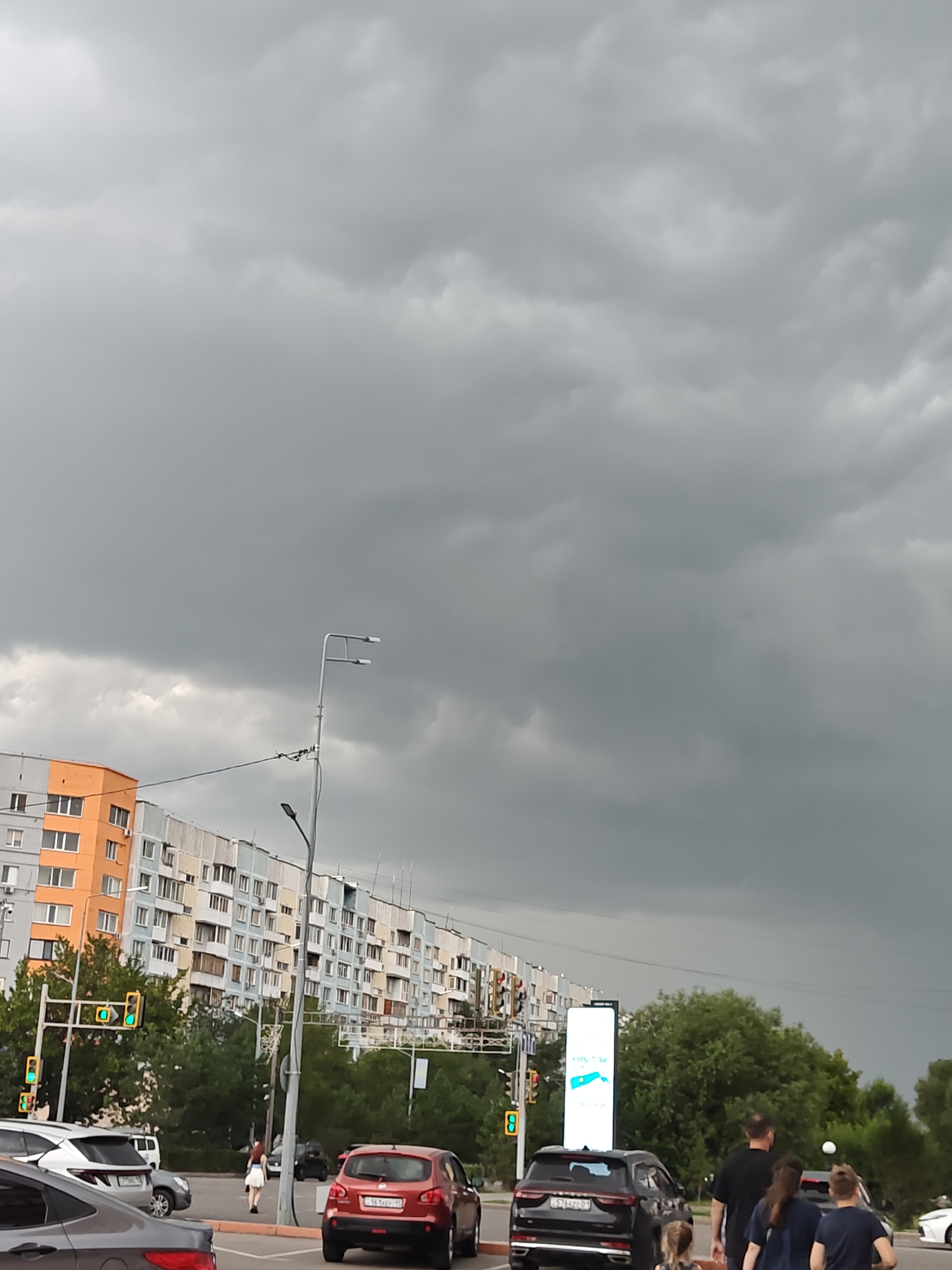 The image depicts a street scene in Petropavl, Kazakhstan, under a heavy, overcast sky. In the foreground, a dark grey car is partially visible on the left, facing away from the camera. To its right, a red SUV is stopped, with its rear facing the camera. Behind the red SUV, a black SUV is also stopped. In the middle ground, a woman in a white dress is walking across the street. Traffic lights are visible, showing green lights. Tall, multi-story apartment buildings line the street, with facades of orange, light blue, and grey. Trees are abundant behind the buildings, and some streetlights are present. In the background, a large digital advertising screen is visible. The overall atmosphere suggests impending rain or a stormy day.