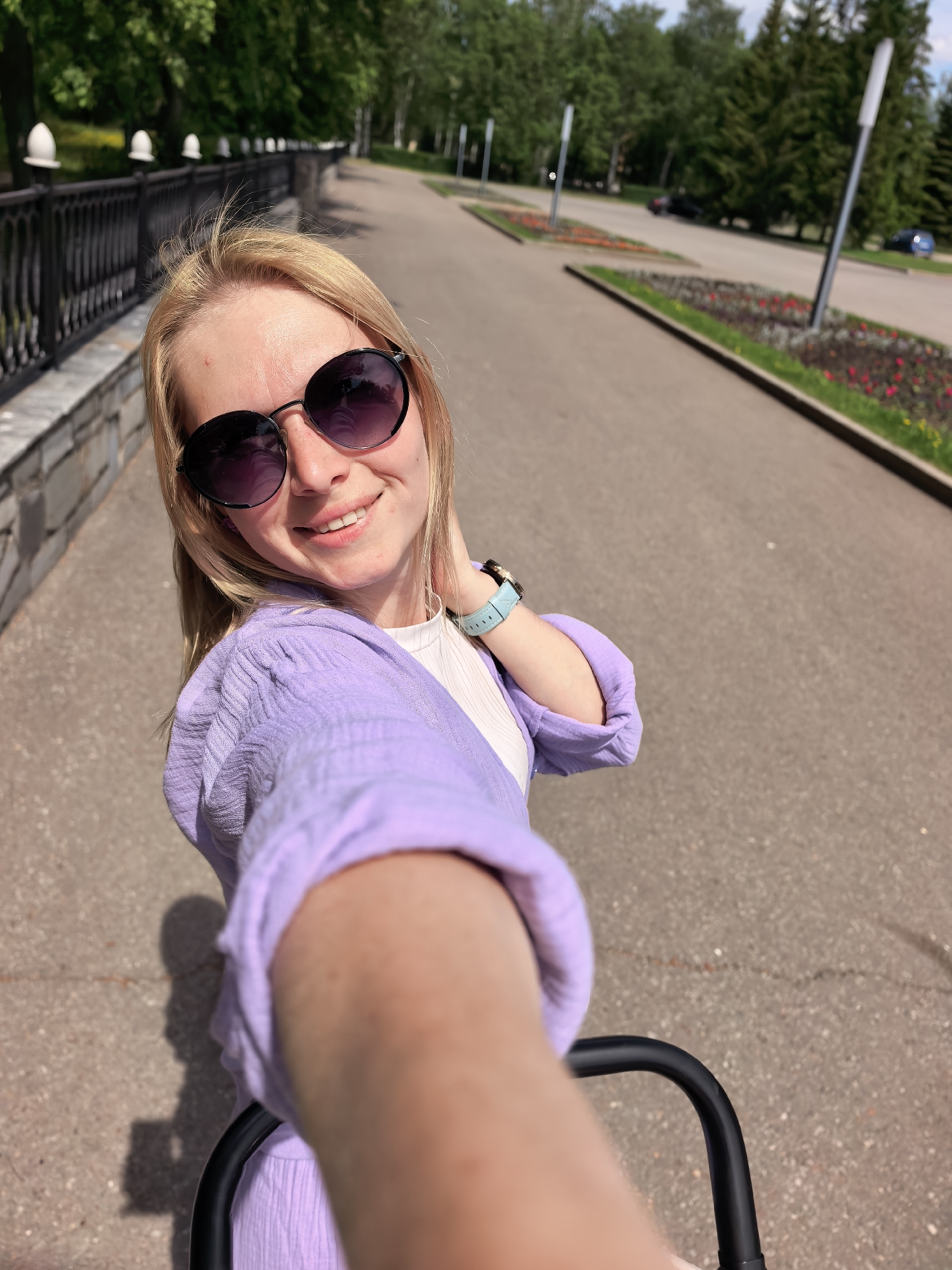 A woman is taking a selfie in a park, likely in Sterlitamak, Russia. She is wearing sunglasses, a purple outfit, and a light blue watch. She is smiling at the camera. Behind her is a paved walkway lined with flower beds and trees. In the background, there is a black fence with decorative caps. She appears to be pushing a stroller.