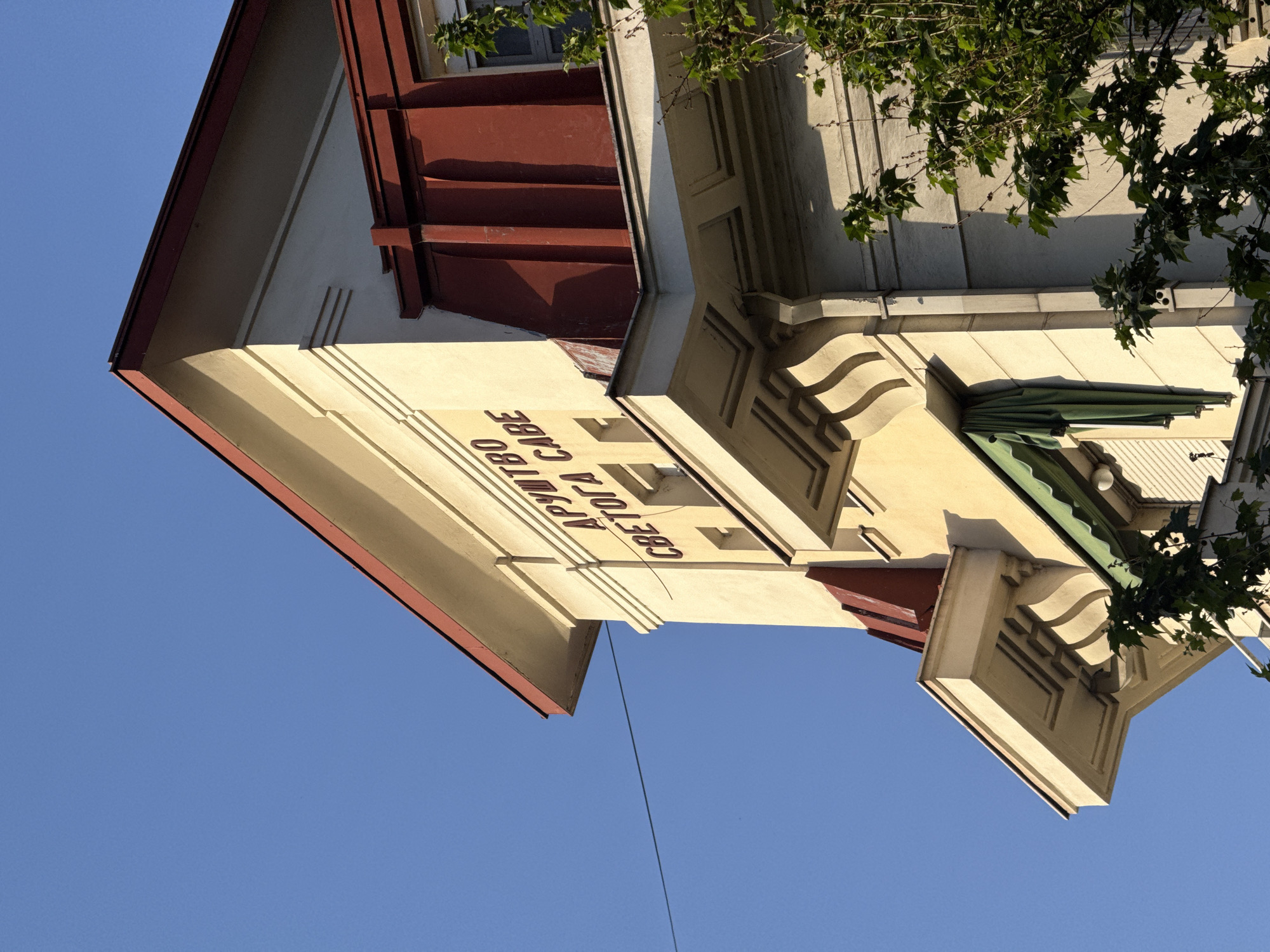 An upward-angled shot features the top of a building against a clear blue sky. The building is light yellow with red trim and intricate architectural details, including moldings and decorative elements. A green awning is partially visible on the right side, and branches with green leaves extend into the frame. Red Cyrillic letters, "АРУШТРО СВЕТОГА САВЕ" are visible on the side of the building. This building is located on Svetog Save Street, Belgrade, Serbia.