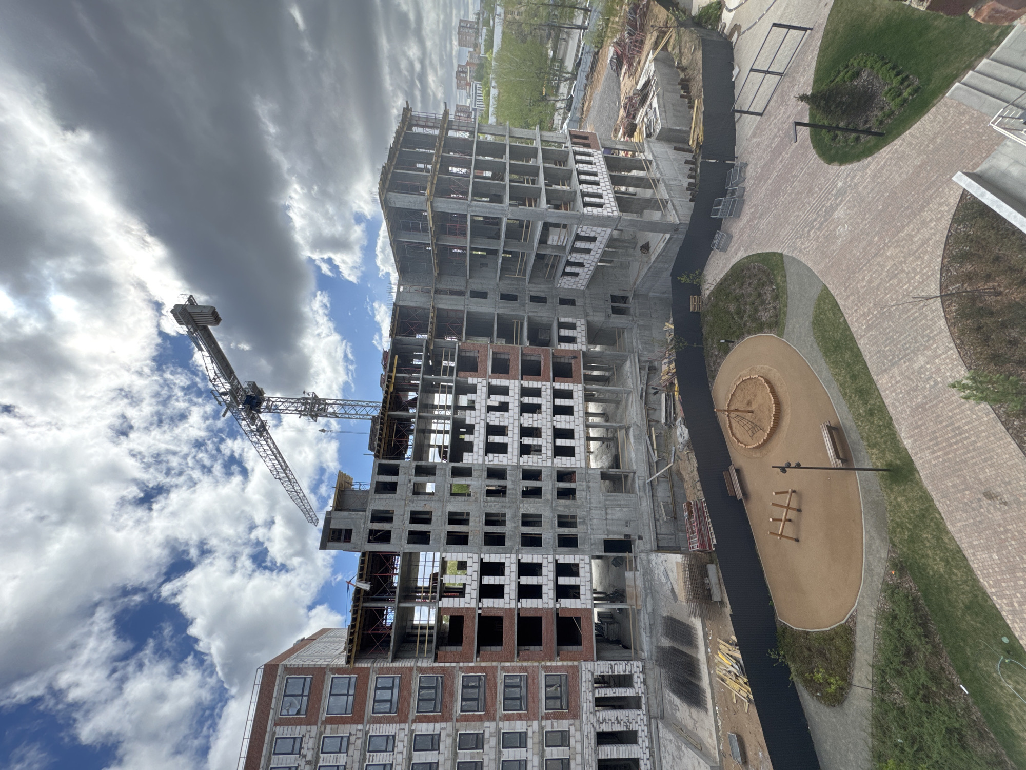 The image depicts a construction site in Люк, Russia. A high-rise building is under construction, with a crane nearby. The building is in its skeletal form, with concrete and brick work visible. A playground with sand, benches, and climbing structures is adjacent to the construction site. The sky is cloudy with patches of blue.
