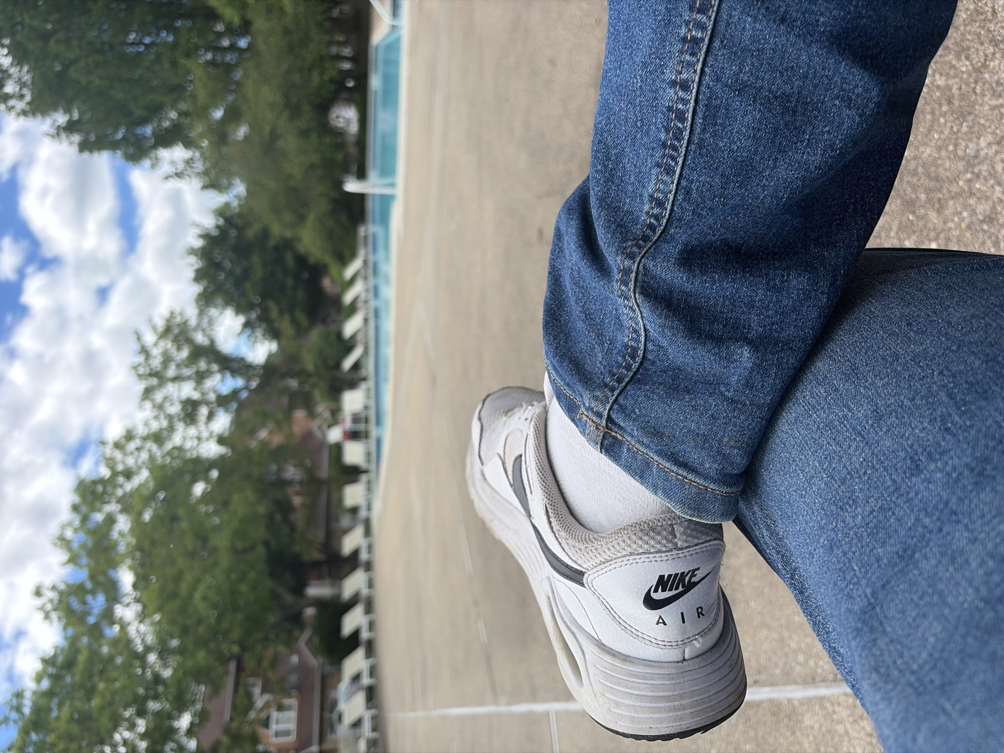 The image shows a person wearing blue jeans and white Nike Air sneakers sitting on a concrete surface. The person has their legs crossed. In the background, there are trees, buildings, and a blue sky with clouds. The location is likely outdoors near a building and greenery, but there is no concrete evidence to support it is within the city of Edison, United States of America. The photo appears to have been taken during daylight hours.