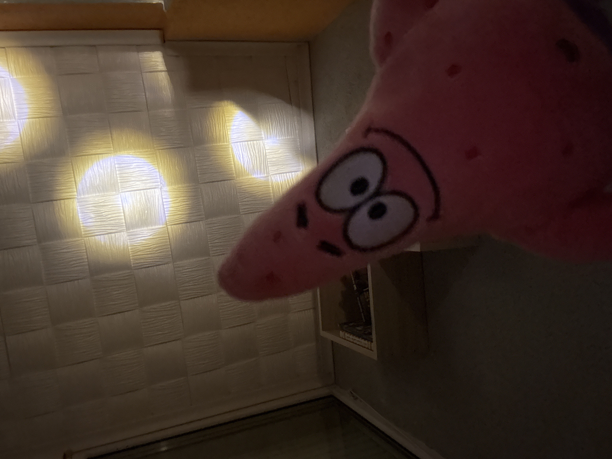 A pink plush toy of Patrick Star from SpongeBob SquarePants dominates the foreground of the image, appearing to look into a dimly lit room. The plush toy is positioned at an angle, with its large, wide-open eyes and stitched smile prominently visible. Its pink, textured surface shows small red dots, characteristic of the character.
The background features an interior room with a distinctive textured wall on the left, possibly covered in square-patterned wallpaper or panels with a ripple-like or woven surface. Several bright, circular spots of yellowish-white light are projected onto this textured wall, creating distinct illuminated areas against the otherwise darker surface. Above the wall, a wooden surface is visible, likely a ceiling or trim.
To the right of the image, behind the plush toy, a dark corner of the room is seen, with part of a recessed shelf or cubbyhole visible on the right wall, containing some indistinct objects. The bottom of the image shows a dark floor or surface with a thin, light-colored trim line. The overall lighting suggests an indoor setting, possibly at night or in a room with limited natural light, relying on artificial illumination. There is no visible text in the image. The scene is set in Panagyurishte, Bulgaria.