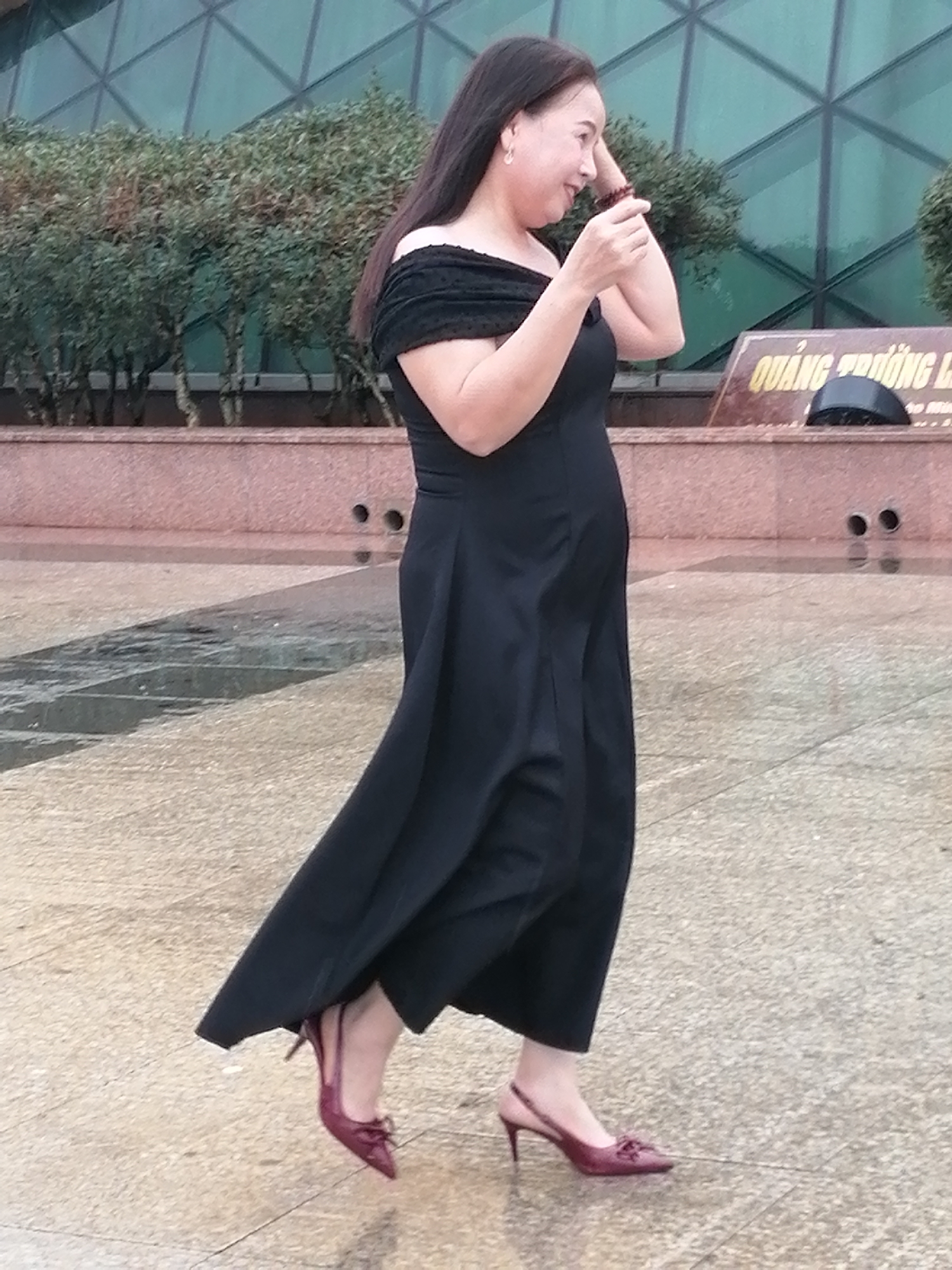 The image depicts a woman wearing a black off-the-shoulder jumpsuit and burgundy heeled shoes. She is standing outdoors on a paved surface, which appears to be wet, suggesting it may have recently rained. In the background, there are trees and a building with a reflective glass facade. A sign with Vietnamese text is partially visible behind the woman. The overall setting suggests an urban or public outdoor space, possibly a plaza or park. Given the sign, the location is likely Da Lat, Vietnam. The woman is looking to her right and appears to be holding a phone or small object in her hand, with her other hand touching her hair.
