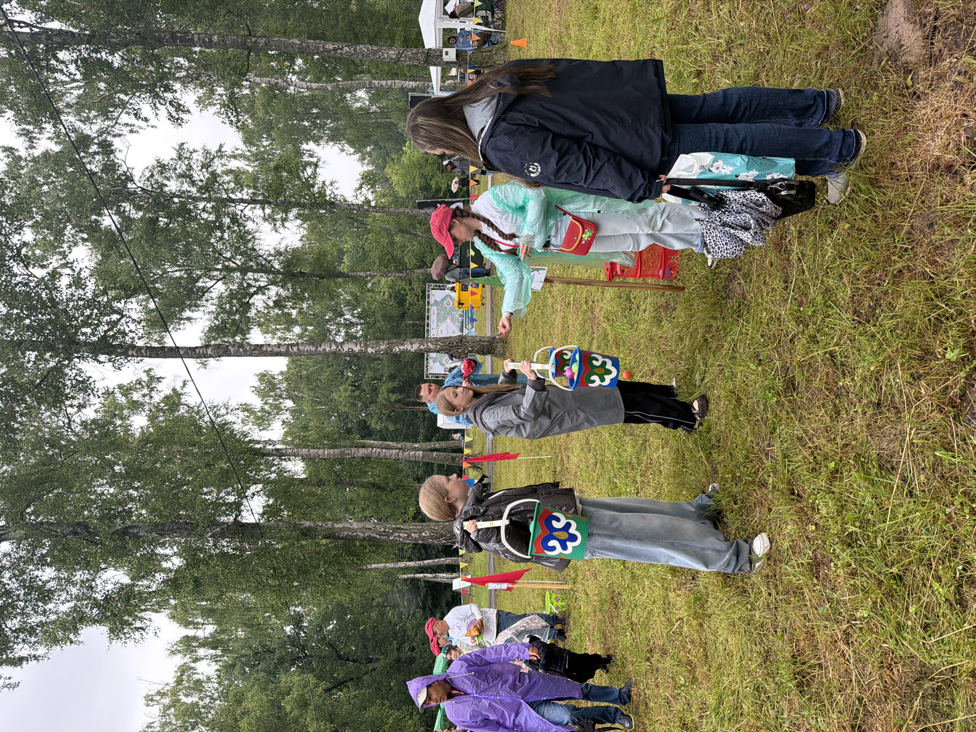 In an outdoor grassy area with numerous birch trees, several individuals are engaged in an activity. The sky is overcast. In the foreground, a young person in a black jacket and light-wash jeans holds a green bucket decorated with colorful floral patterns. To their right, another young person in a grey hooded coat and black pants holds a similar bucket, also adorned with floral designs. To the left, a person in a purple hooded raincoat walks away from the camera, carrying a black backpack. Further back, a woman in a turquoise rain poncho and a pink cap is interacting with others, holding a small red purse. In the background, tents, people, and colorful banners are visible, suggesting an event or festival. The ground is covered in green grass, with some patches of dry grass and scattered debris. Red flags on poles are placed sporadically throughout the area.