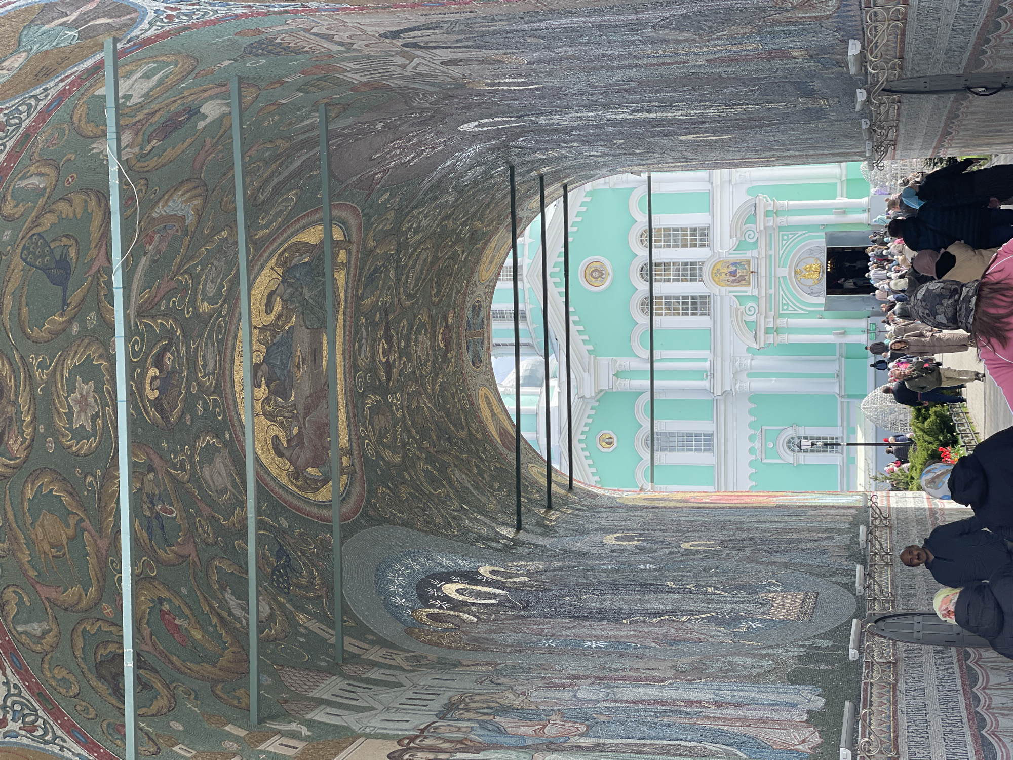 Here's a detailed description of the image:
**Scene:**
The image depicts a view through a mosaic-covered archway, leading to an outdoor area where a light green and white building stands. A crowd of people gathers near the entrance of the building.
**Architecture:**
The archway is extensively decorated with mosaic artwork featuring religious imagery. The building in the background has a distinctive architectural style, characterized by its pastel color scheme, arched windows, and decorative elements.
**People:**
The crowd consists of people of diverse ages and appearances. Some are standing near the entrance, possibly waiting to enter, while others are walking around. Their attire suggests an outdoor setting.
**Objects:**
Beyond the archway, there are hints of greenery and other outdoor elements, suggesting a courtyard or park-like setting. There are metal bars or supports within the archway structure.