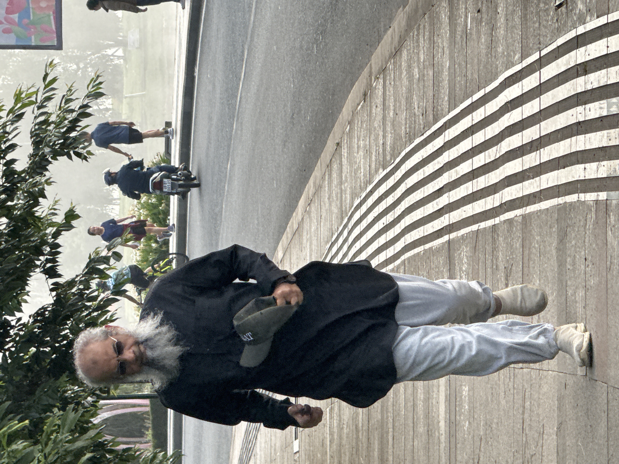 The image depicts a sidewalk in Da Lat, Vietnam, with a man walking towards the camera. He has a long grey beard and is wearing sunglasses, a black long-sleeved shirt, and light grey sweatpants. He is holding a dark green cap in his right hand. The sidewalk has raised tactile paving strips. In the background, a paved road is visible with a motorcycle and several pedestrians. Trees with dense foliage overhang the sidewalk. The overall lighting suggests it is daytime.