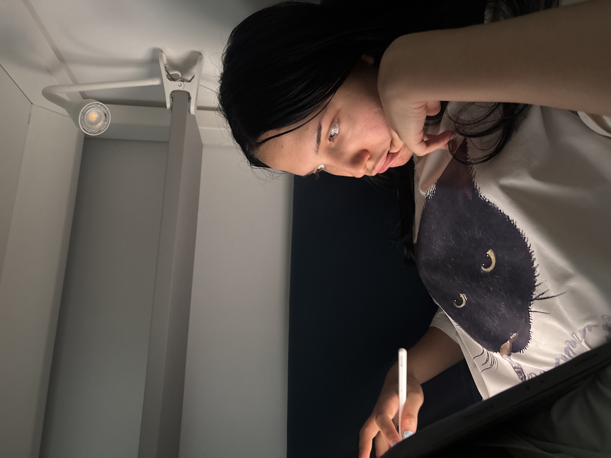 The image shows a young person lying down, holding a stylus to a tablet. They are wearing a white t-shirt with a black cat illustration. A clip-on lamp is attached to a shelf above them. The background consists of a dark blue surface and light grey shelves. The scene is dimly lit, with light emanating from the tablet and the clip-on lamp. The person's face is illuminated from the side, and they are looking intently at the tablet. The overall impression is one of focused activity in a cozy, private space, likely a bedroom or a study area. There is no indication that this image is located in Cupra Marittima, Italy.