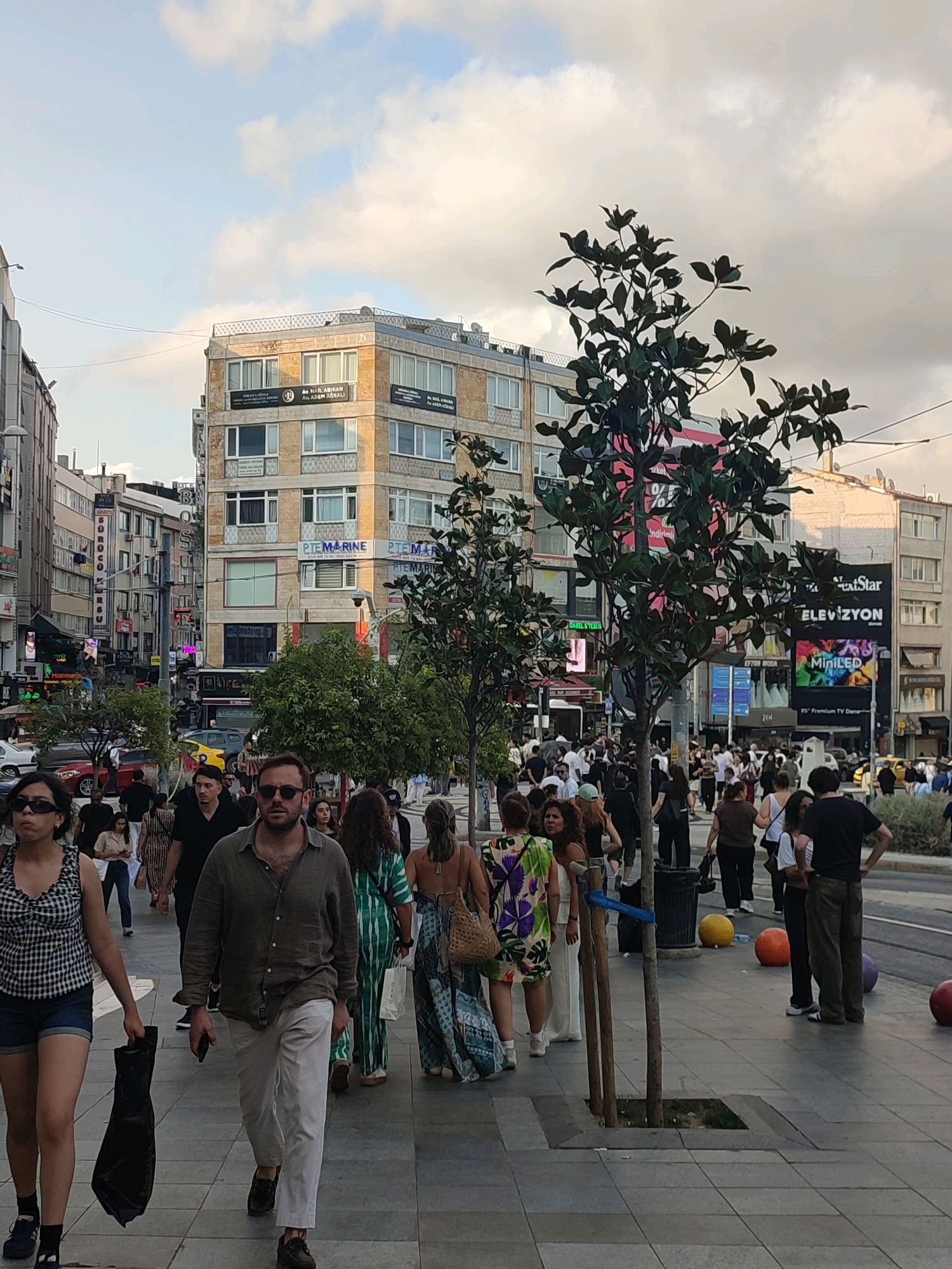 In Kadıköy, Turkey, people walk along a bustling street lined with buildings. In the foreground, a man in sunglasses and a linen shirt walks towards the camera, holding a phone. To his left, a woman in a checkered top and shorts carries a black bag. Behind them, a crowd of people are walking and standing. Buildings with advertisements and windows dominate the background. Several trees are planted along the sidewalk. The sky above is cloudy.