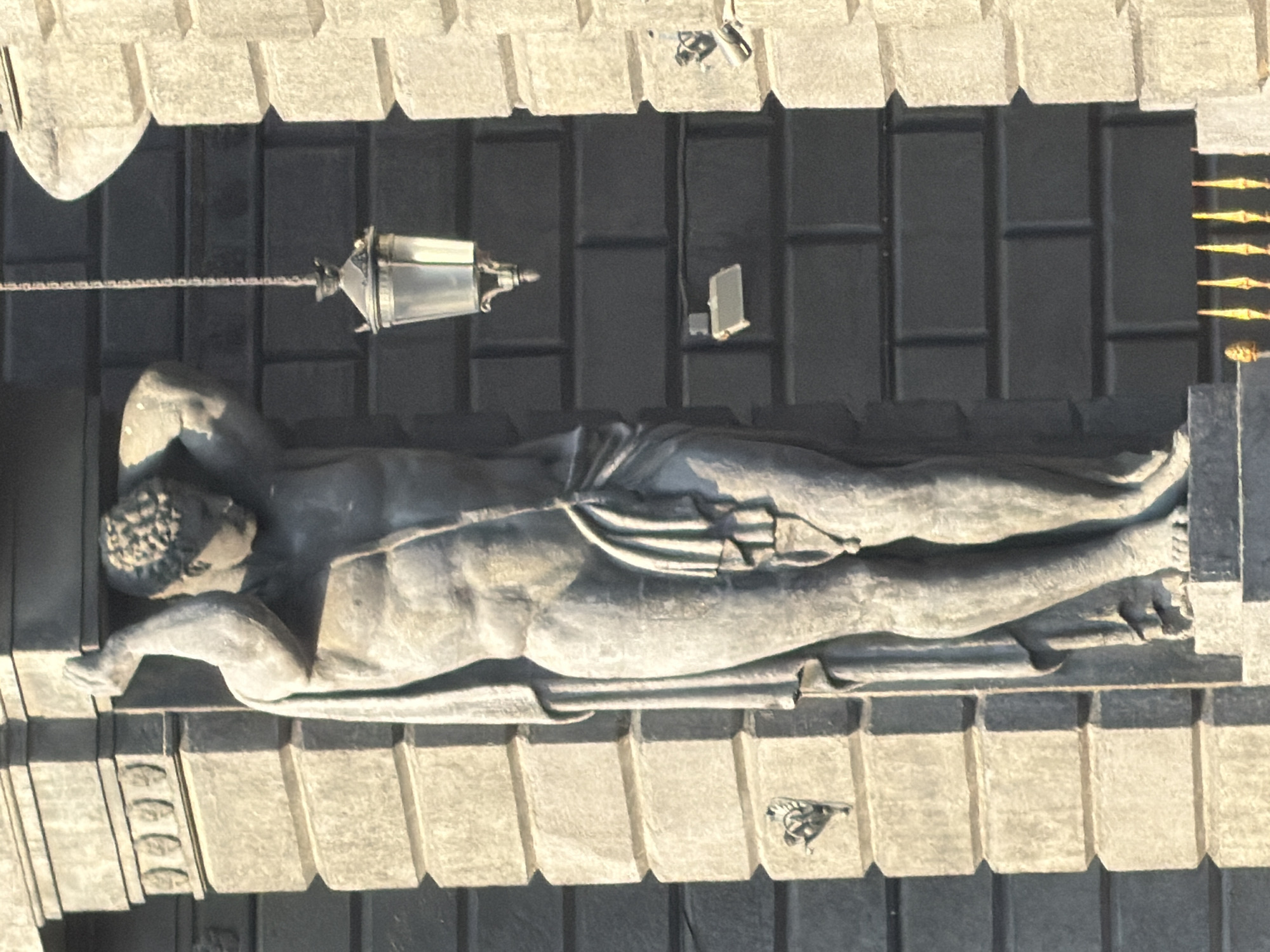 The image shows a sculpture of a reclining male figure, likely made of stone or concrete. The sculpture is located above a dark brick facade, possibly part of a building. A hanging lamp with a chain is positioned near the sculpture's upper body. The setting appears to be an urban environment, possibly in Saint Petersburg, Russia. Architectural details suggest a classical or neoclassical style, characteristic of Saint Petersburg's historic buildings. The image is taken from an elevated perspective, looking down on the scene.