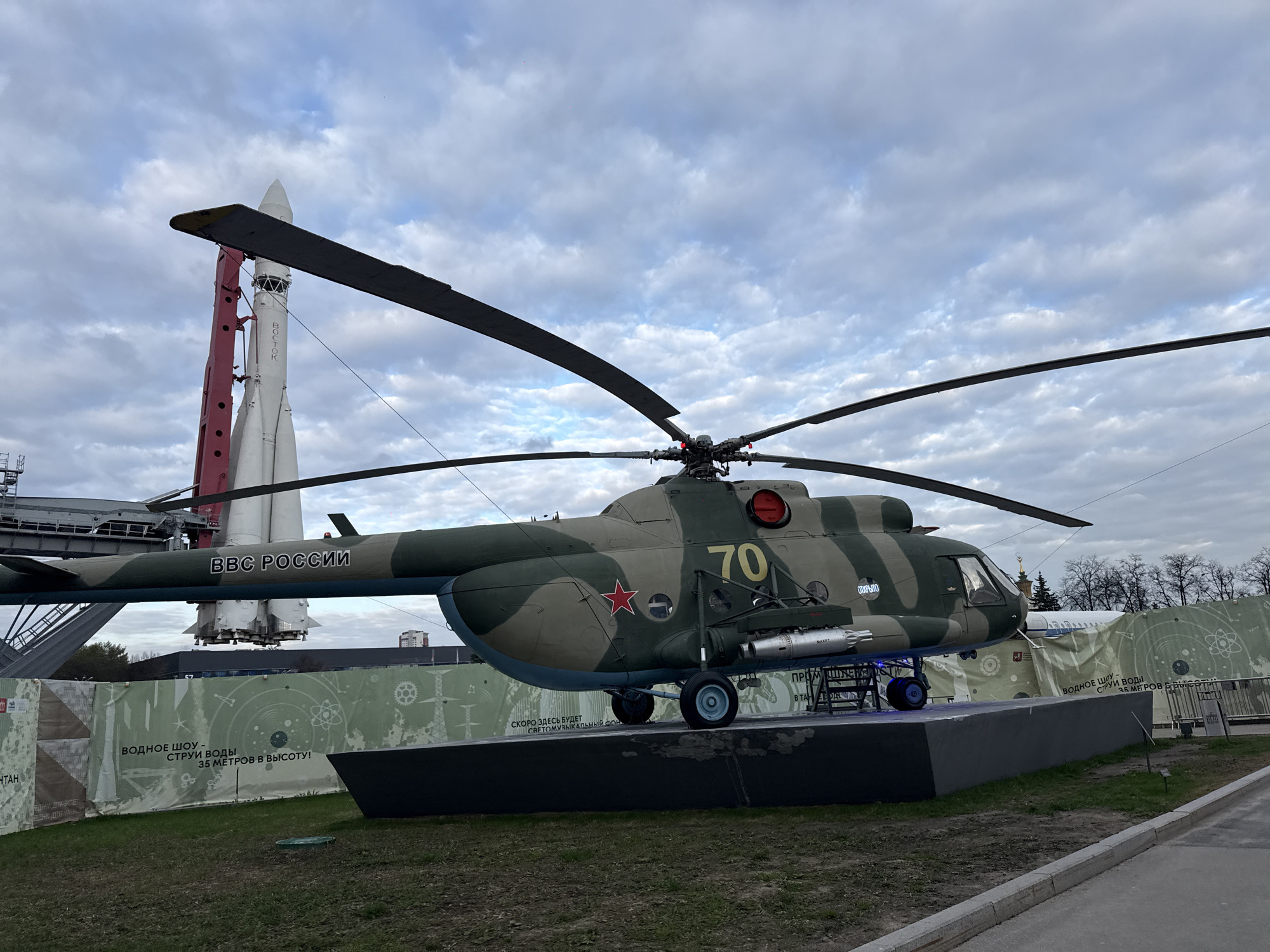 The image captures a scene at VDNH (Exhibition of Achievements of National Economy) in Moscow, Russia. The foreground features a Mil Mi-24 Hind helicopter on display. The helicopter is painted in a camouflage pattern with the number "70" and the inscription "BBC Poccuu" ("VVS Rossii" in English: "Russian Air Force"). The helicopter sits atop a dark, elevated platform. Behind the helicopter is a Vostok rocket. A banner displaying Russian text and diagrams relating to a water show, and science-themed graphics add to the background context. The sky is overcast with gray clouds. The ground is covered in grass and there is a paved pathway in the lower right corner of the image. Bare trees are visible in the distance to the right of the helicopter.
