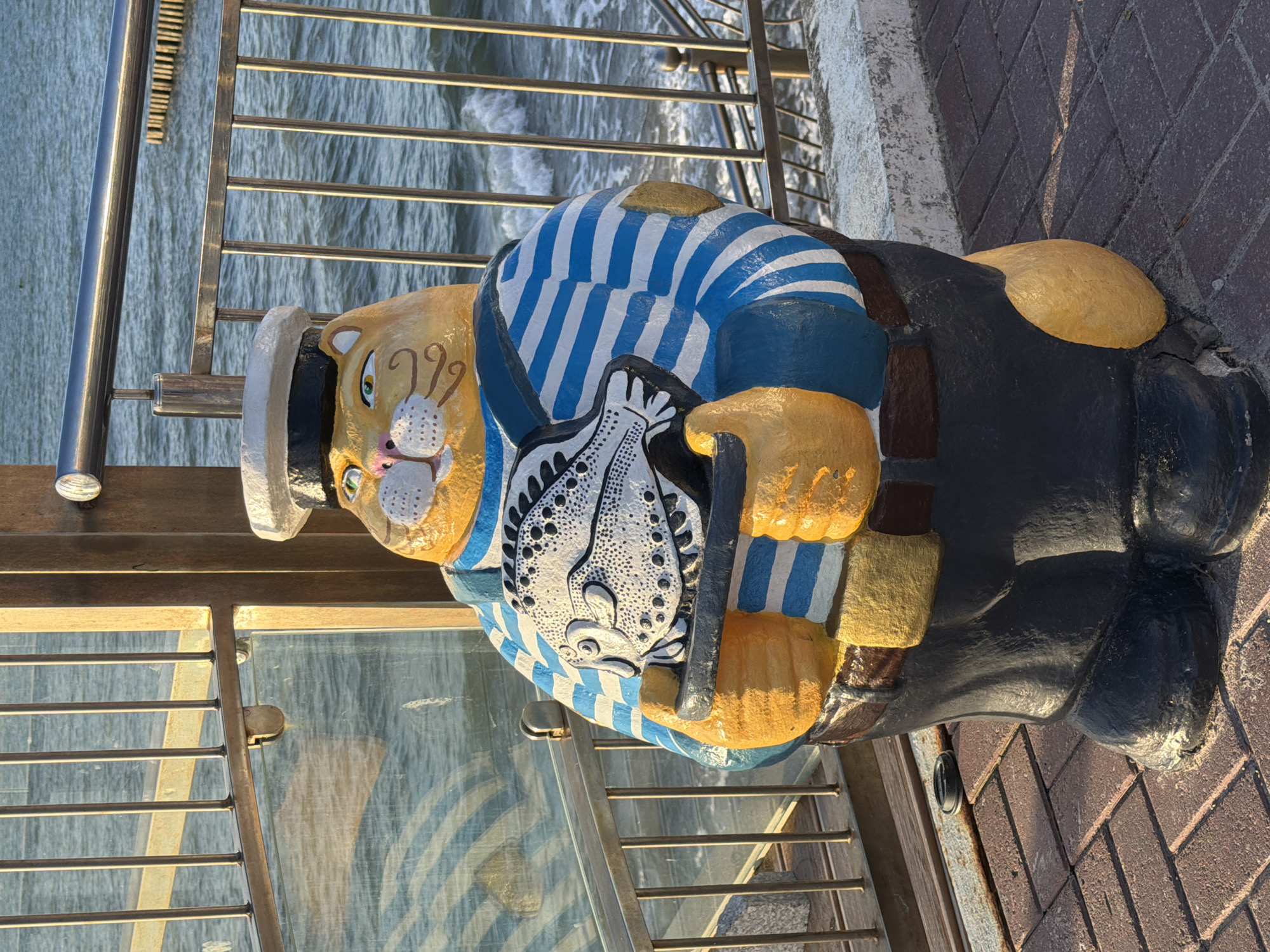 The image shows a sculpture of a plump, anthropomorphic cat dressed as a sailor. The cat is wearing a white sailor's cap with a black band, a blue and white striped shirt with overalls, and dark pants. It is holding a stylized fish in its paws. The sculpture is yellow and orange with painted details on its face, including green eyes and whiskers. It is standing on a brick pathway next to a metal railing that overlooks a body of water with gentle waves. The background shows the sea and a wooden structure. This sculpture is located in the city of Zelenogradsk, Russia, likely on the promenade or a public space near the coast.