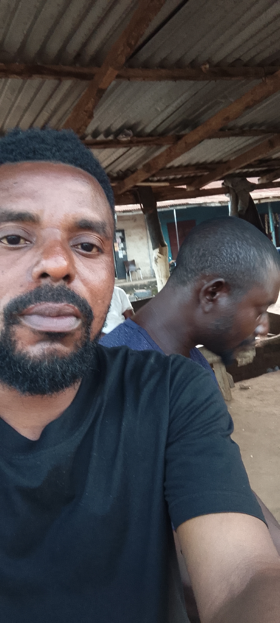 The image is a selfie taken in what appears to be an outdoor, rustic setting, possibly in Gboko, Nigeria. The foreground features the face of a man with dark skin, a goatee, and a black t-shirt, looking directly at the camera. His left eye is visible, as is his nose and mouth. Behind him and to his right, another man's head and shoulder are visible, with his face turned away from the camera. This second man has short, dark hair and is wearing a blue shirt. In the background, there is a corrugated metal roof supported by wooden beams. Further back, a building with windows and what looks like a chair and some shelves are visible. The ground is a dirt or sand surface. The overall environment suggests a casual, informal gathering or a local roadside establishment.