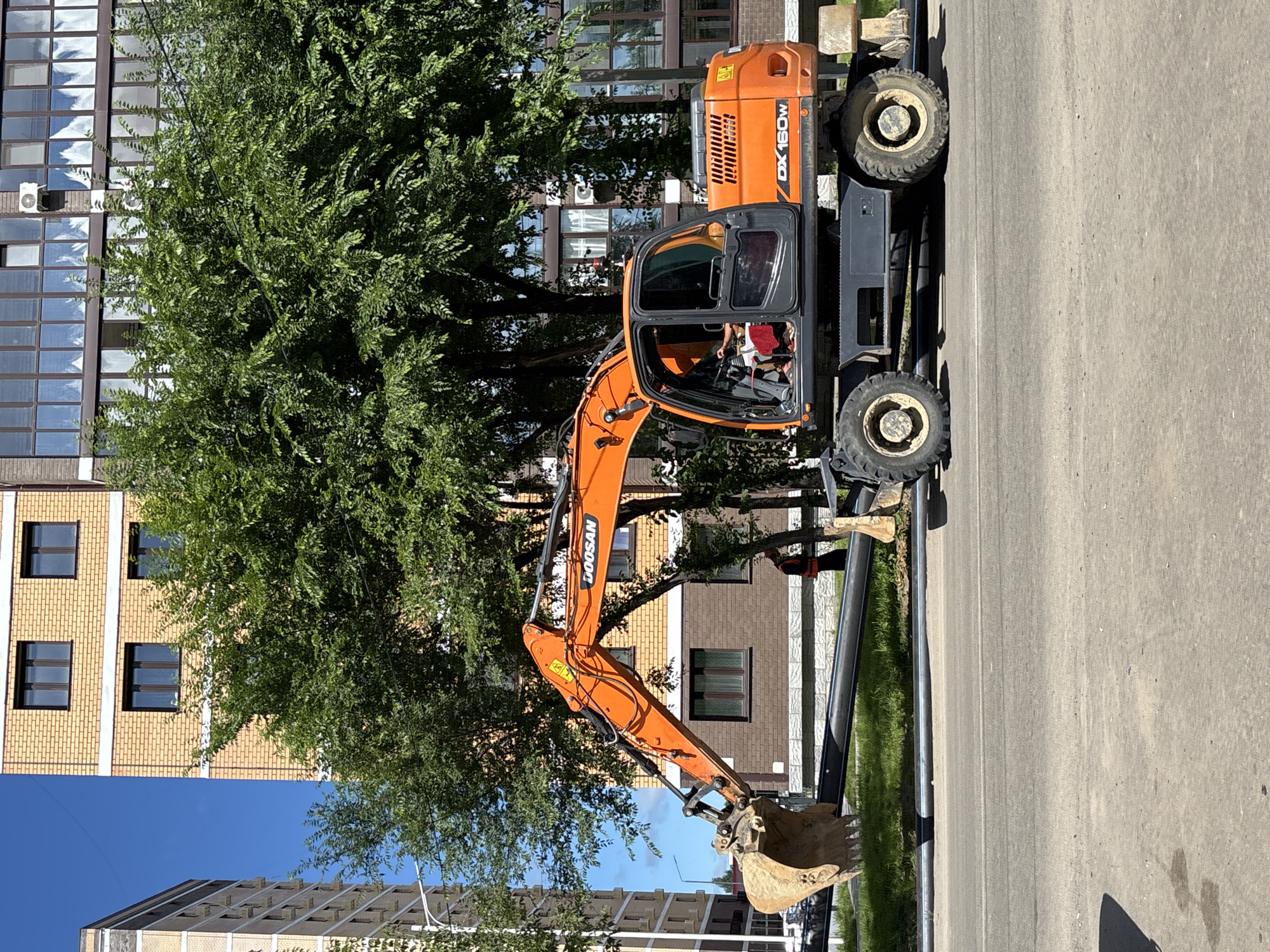 An orange Doosan DX160W wheeled excavator is parked on the side of a paved road, partially obscuring a large black pipe lying on the grass. The excavator's boom is raised, and its bucket is resting on the ground near the pipe. A person wearing a high-visibility vest is visible in the background, standing near a brown brick building. Behind the excavator, a modern apartment building with yellow brick facade and numerous windows is seen, alongside a tall building with a grey facade. Lush green trees are scattered throughout the scene, with a clear blue sky overhead. The image appears to have been taken in the city of Blagoveshchensk, Russia, likely during daytime.