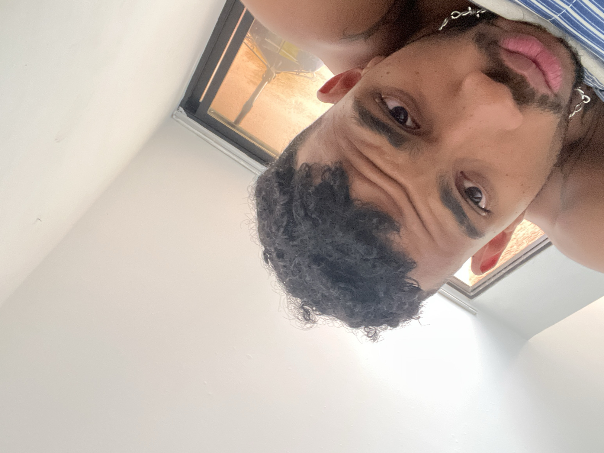 The image shows a close-up, tilted selfie of a young man lying down in what appears to be a bedroom. The man has dark curly hair and is looking towards the camera with his eyes open. He is wearing a silver chain necklace. A portion of a window with orange-tinted glass is visible in the background, suggesting it might be daytime. The man is lying on a striped blue and white surface, possibly a pillow or bedding. The room has white walls and a white ceiling. The orientation of the image suggests it was taken from a low angle. The overall impression is an informal, candid shot. There is no specific information in the image to link it to Porlamar, Venezuela.