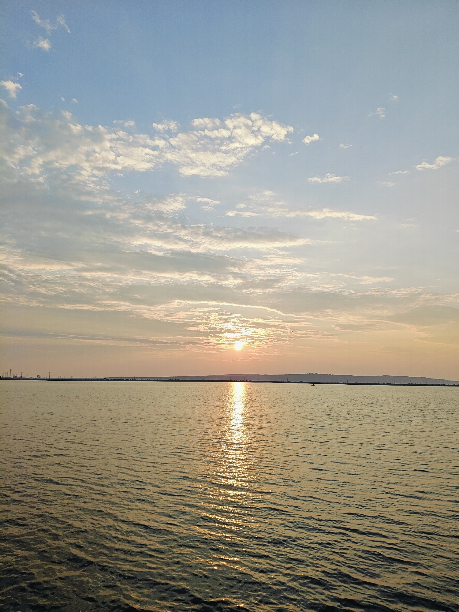 The image depicts a tranquil, wide body of water under a sky with scattered clouds and a setting sun. The water surface is calm with gentle ripples, reflecting the warm, golden hues of the sunset. A distant horizon line separates the water from a landmass that appears to be a low-lying shore, dotted with what look like industrial structures and possibly wind turbines along its length. The sky transitions from a soft blue at the top to warmer tones of orange and yellow near the horizon, where the sun is partially obscured by clouds. The overall impression is one of peacefulness and natural beauty, enhanced by the soft lighting of dusk. The scene is likely a view of the Kama River or one of its reservoirs, given the location in Chaikovsky, Russia, a city known for its industrial development and proximity to the river.