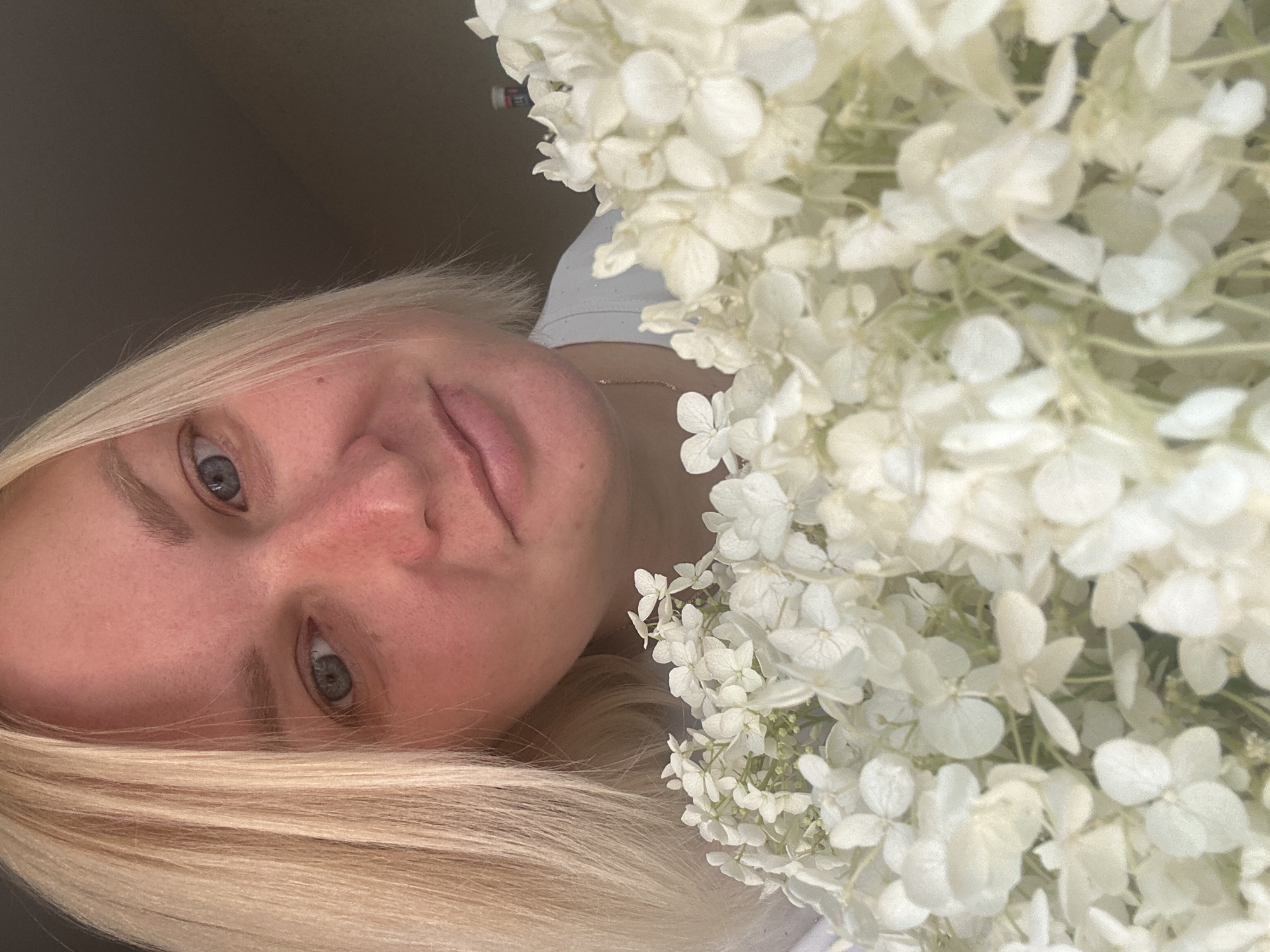 A blonde woman with blue eyes holds a bouquet of white hydrangeas in front of her face, obscuring most of her lower face and body. She is wearing a white t-shirt and a thin gold chain necklace. The background is dark and out of focus, suggesting an indoor setting. The image is a close-up, portrait-style photograph. There are no discernible elements of the city of Chaikovsky, Russia, present in the image.