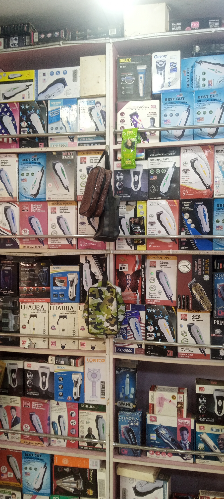 The image shows a retail display of hair clippers and grooming products on shelves. The products are mostly packaged in colorful boxes with images of the clippers and brand names. There are no people visible in the image. The scene appears to be a small store or market stall. The items are arranged in an orderly fashion on multiple shelves, suggesting a commercial setting. The lighting is bright, likely from overhead store lights. The overall impression is one of a well-stocked retail environment selling personal grooming appliances. It is not possible to determine the specific location within Jalingo, Nigeria, based on the visual information provided.