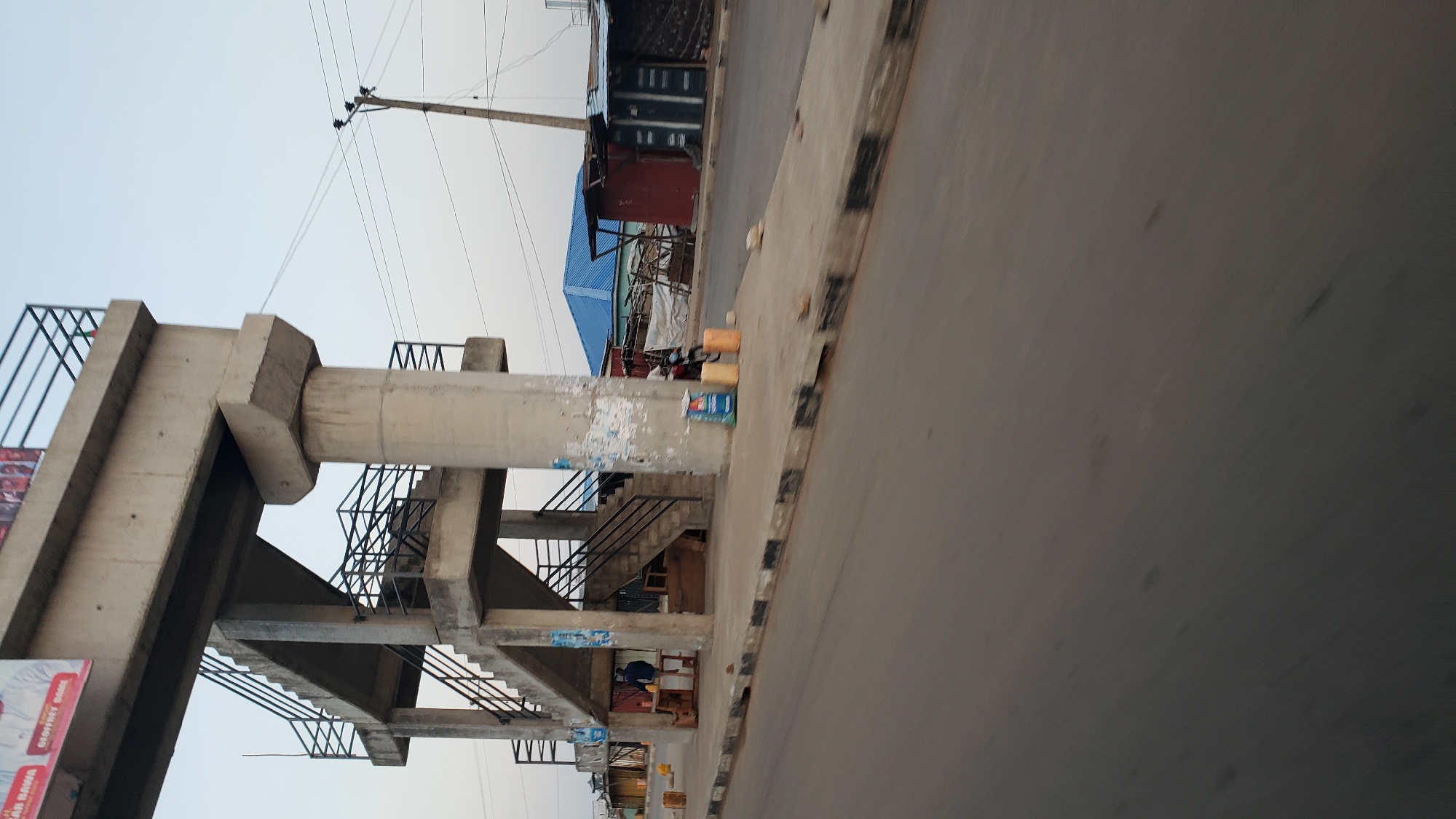 The image shows an outdoor scene in Jalingo, Nigeria, at what appears to be dusk or early morning with a clear sky. In the foreground, a paved road with a distinctive black and white striped curb runs diagonally from the bottom left to the top right. To the right of the road is a sidewalk.
Dominating the left side of the image is a concrete pedestrian overpass with a series of staircases and landings, supported by thick concrete pillars. Black metal railings line the stairs and the walkway at the top. A large banner, partially visible at the top left of the overpass, features text and images.
On the sidewalk beneath the overpass, there are various objects. A man in a dark jacket is standing near a small wooden stall or kiosk. Several yellow jerrycans are placed on the ground, along with blue containers, and some wooden planks. A motorcycle is parked near the blue containers. Further back, past the overpass supports, a series of structures are visible, including buildings with blue and red roofs, and what appear to be market stalls or shops. Power lines and a utility pole are visible in the background, extending across the sky. The overall impression is a busy, possibly commercial area in an urban setting.