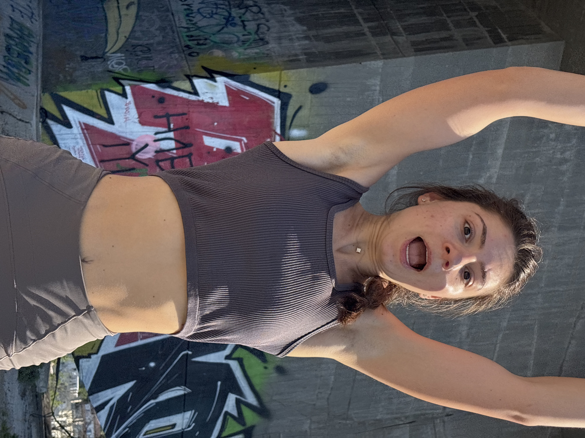 A young woman with brown hair and an open mouth is photographed from the chest up, arms raised above her head. She is wearing a ribbed, taupe-colored crop top and matching leggings. Her skin is tanned, and she has a small, square diamond necklace. The background is a concrete wall covered in graffiti. The image was taken outdoors, likely in an urban setting.