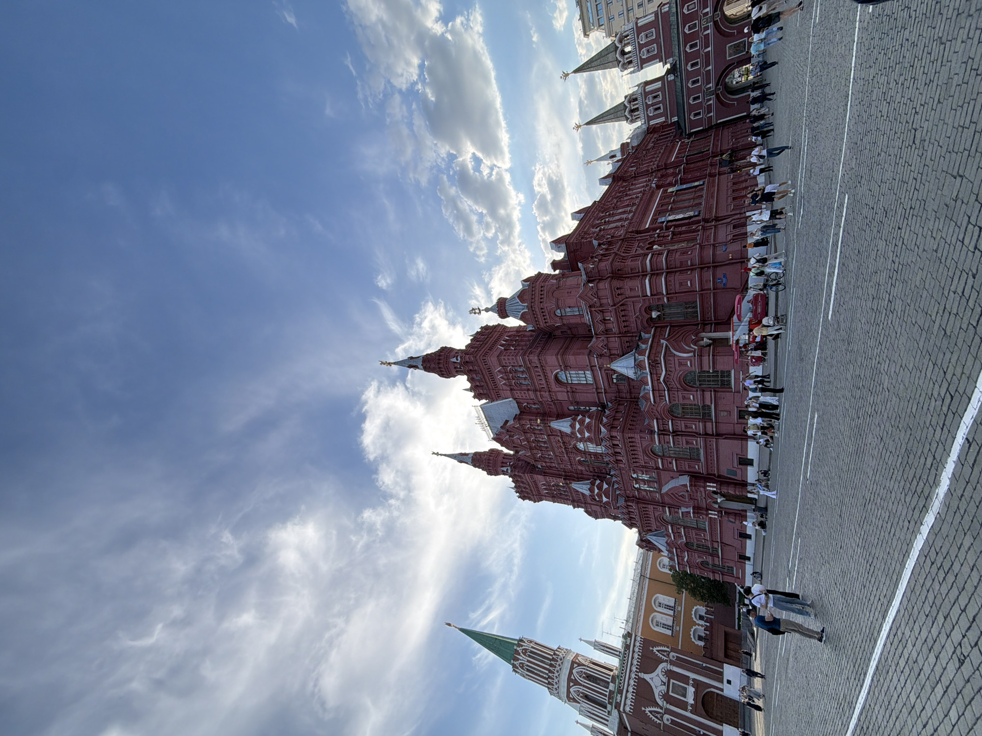 The image shows the State Historical Museum in Moscow, Russia, alongside part of Red Square. The red brick building has multiple spires and is topped with double-headed eagles. People are walking on the cobblestone square. The sky is blue with scattered clouds.