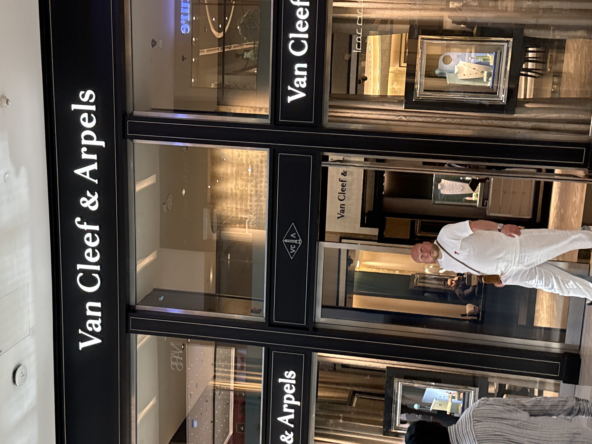 A man wearing a white t-shirt and white pants walks by a Van Cleef & Arpels store, likely located in Dubai, United Arab Emirates, as indicated by the Arabic script visible on the store's exterior. The man has a brown satchel slung across his chest. Another person, partially visible on the left, is wearing a striped shirt. Inside the store, jewelry displays are visible through the large windows. The store's name, "Van Cleef & Arpels," is prominently displayed in white lettering on a black background above the entrance. A logo featuring "VC A" within a diamond shape is also visible. The store appears to be in a well-lit indoor shopping environment, possibly a mall.