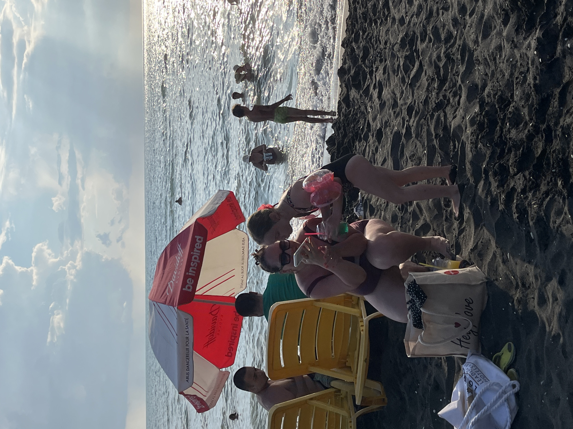 The image depicts a beach scene in Ureki, Georgia, with several people enjoying the water and the sand. In the foreground, a woman is crouched down, looking at her smartphone, while a young girl stands next to her, holding a drink with a straw and wearing arm floaties. Behind them, two yellow lounge chairs are visible, and further back, several people are in the ocean. A red and white beach umbrella is open, providing shade. The beach has dark sand, and the water is calm with sunlight reflecting off the surface. There are two beach bags on the sand near the woman and girl. In the background, there are clouds in the sky, suggesting it may be late afternoon or early evening.