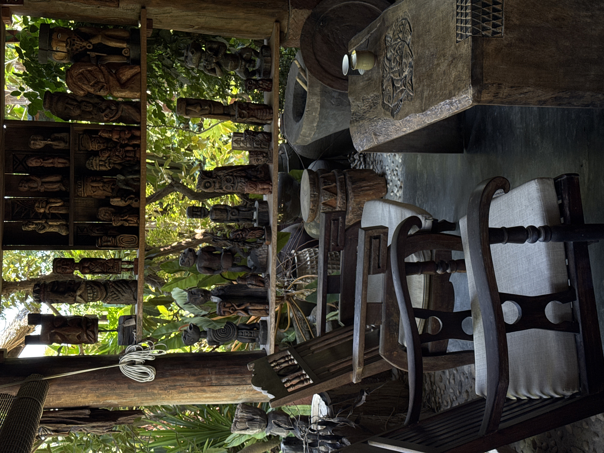 The image shows an indoor space with a collection of wooden carvings and artifacts, likely in Đà Nẵng, Vietnam, given the artifacts' style. The space contains wooden chairs with light-colored cushions, a large wooden structure carved with intricate designs, and numerous carved wooden figures displayed on shelves. The view extends outwards to lush greenery, implying a garden or outdoor setting. A coiled wire hangs from a wooden beam near the carved figures.