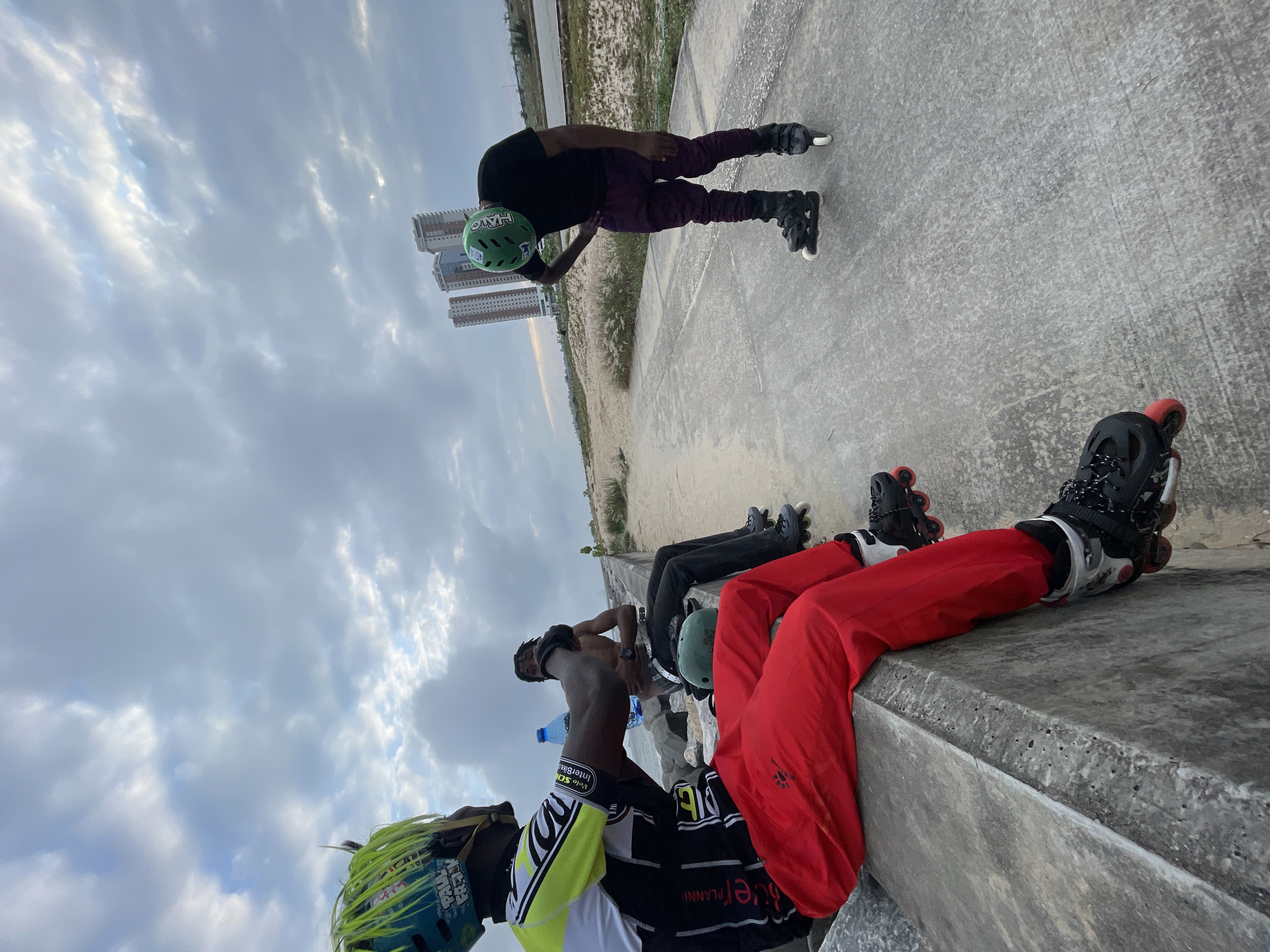 In Ilado, Nigeria, three individuals are present in a coastal recreational area. The sky above is filled with dramatic, overcast clouds, suggesting an impending weather change or a recent storm. Two individuals are seated on a concrete barrier. The person on the left, facing away from the camera, is wearing a black and neon yellow cycling jersey with "PLANNY" written on the back, red pants, and black rollerblades with red accents. They are holding a blue water bottle. Beside them, another person is seated, wearing black pants and black rollerblades. A green helmet rests on the ground in front of them. The third individual is standing on the paved path in front of the concrete barrier, wearing a black t-shirt, purple pants, and black rollerblades. They are wearing a green helmet and have one hand on their hip, appearing to be looking down. In the background, several tall buildings are visible, along with sparse vegetation and the ocean. The overall scene suggests a pause in an outdoor activity, likely rollerblading, due to the weather or a break.