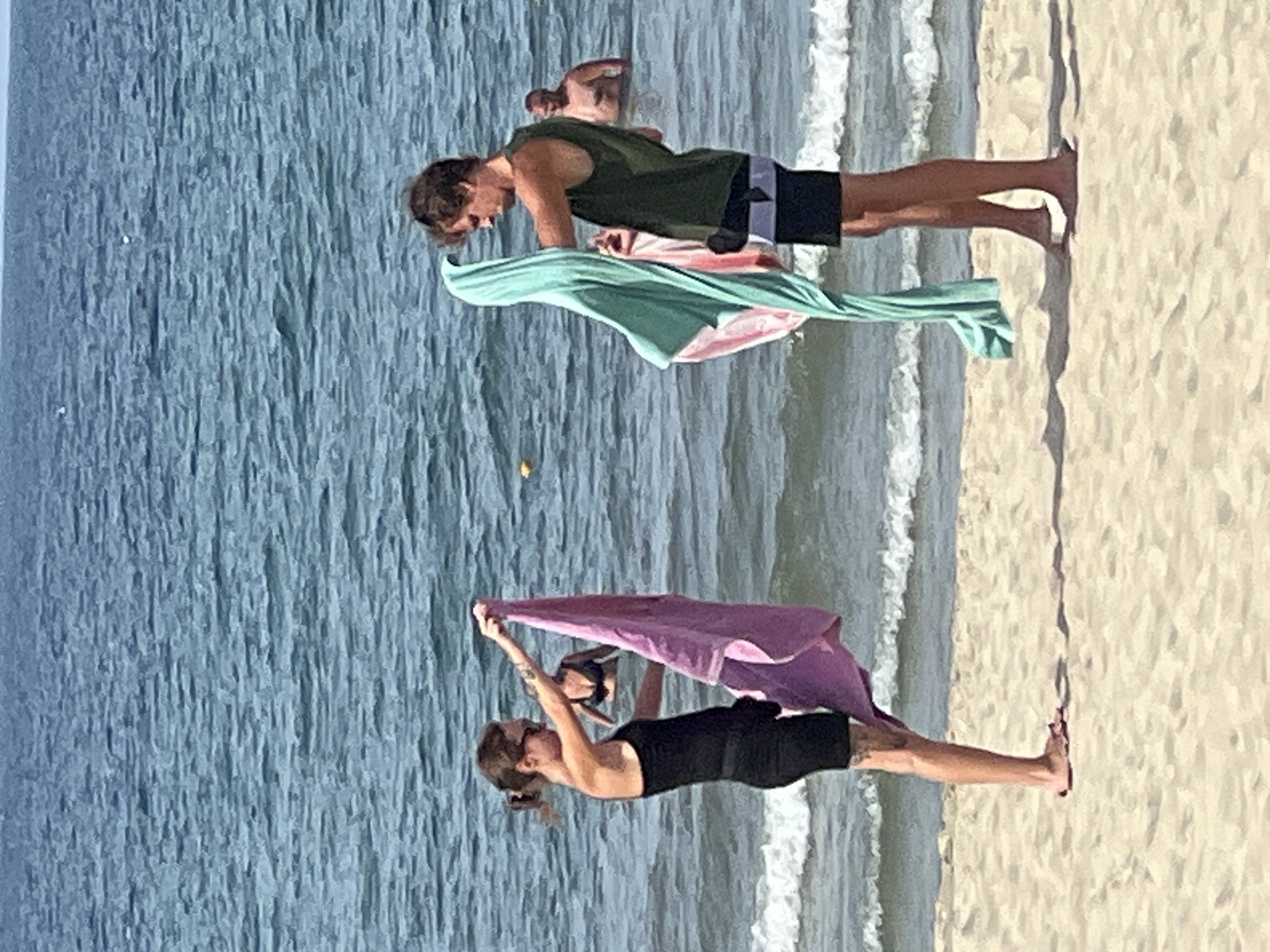 The image captures a beach scene with two individuals holding towels. The woman on the left wears a black dress and sunglasses and holds a purple towel. The man on the right wears a green sleeveless shirt and black shorts and holds a light blue and pink towel. Both are standing on a sandy beach with the ocean in the background. Waves are visible, and there are other people in the water, including a man swimming and a woman in a bikini. The location is identified as a beach in Đà Nẵng, Vietnam.
