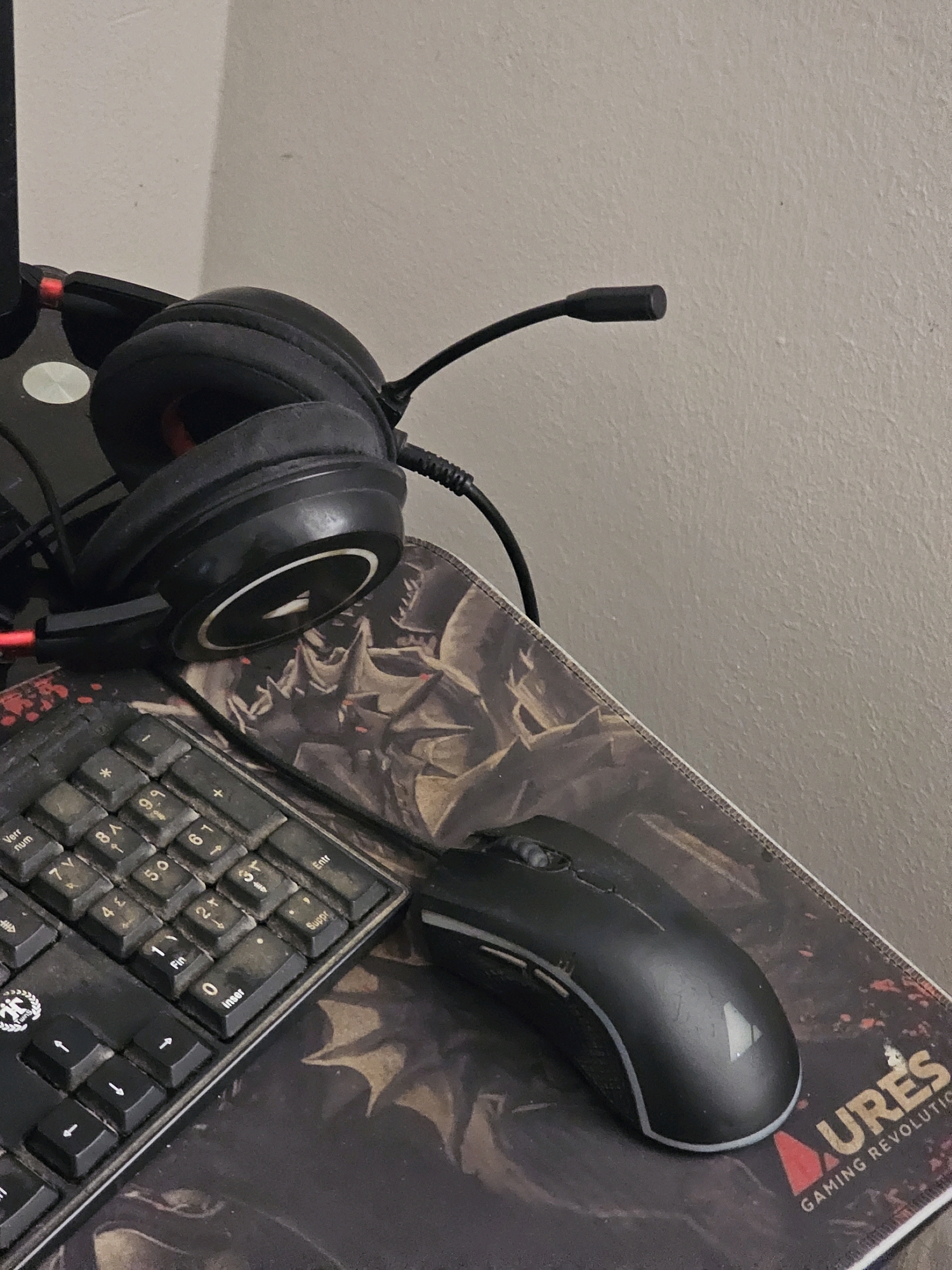 The image displays a close-up, slightly angled shot of a computer desk setup. Dominating the foreground is a black gaming mouse, resting on a mousepad that features a dark, fantasy-themed graphic with the partial text "URES GAMING REVOLUTIO" visible. To the left, a portion of a black keyboard is visible, showing some keys with alphanumeric characters and symbols. Above the keyboard and mousepad, a pair of black gaming headphones with red accents are positioned, their microphone boom extending towards the right. The background consists of a plain, light-colored wall, suggesting an indoor setting. There is no discernible indication of the city of Batna, Algeria.