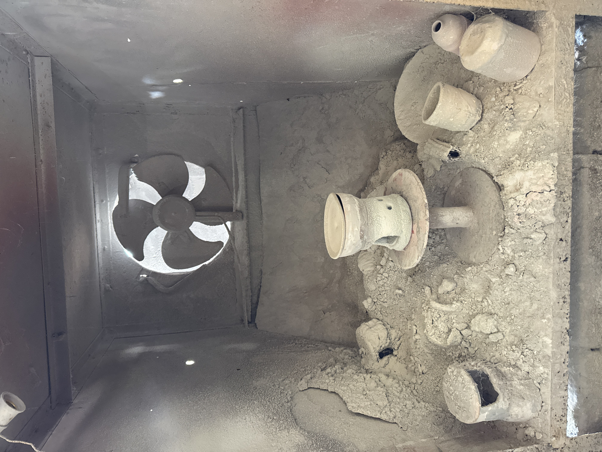 The image shows the interior of a kiln, possibly located in Kediri, Indonesia. On the left is a large fan, likely for ventilation or temperature regulation. The right side of the kiln contains several ceramic pieces covered in white dust or powder, including pots, cylinders, and other formed clay objects. The environment is dusty and appears to be an industrial setting. The kiln is box-like in shape. No people are visible.