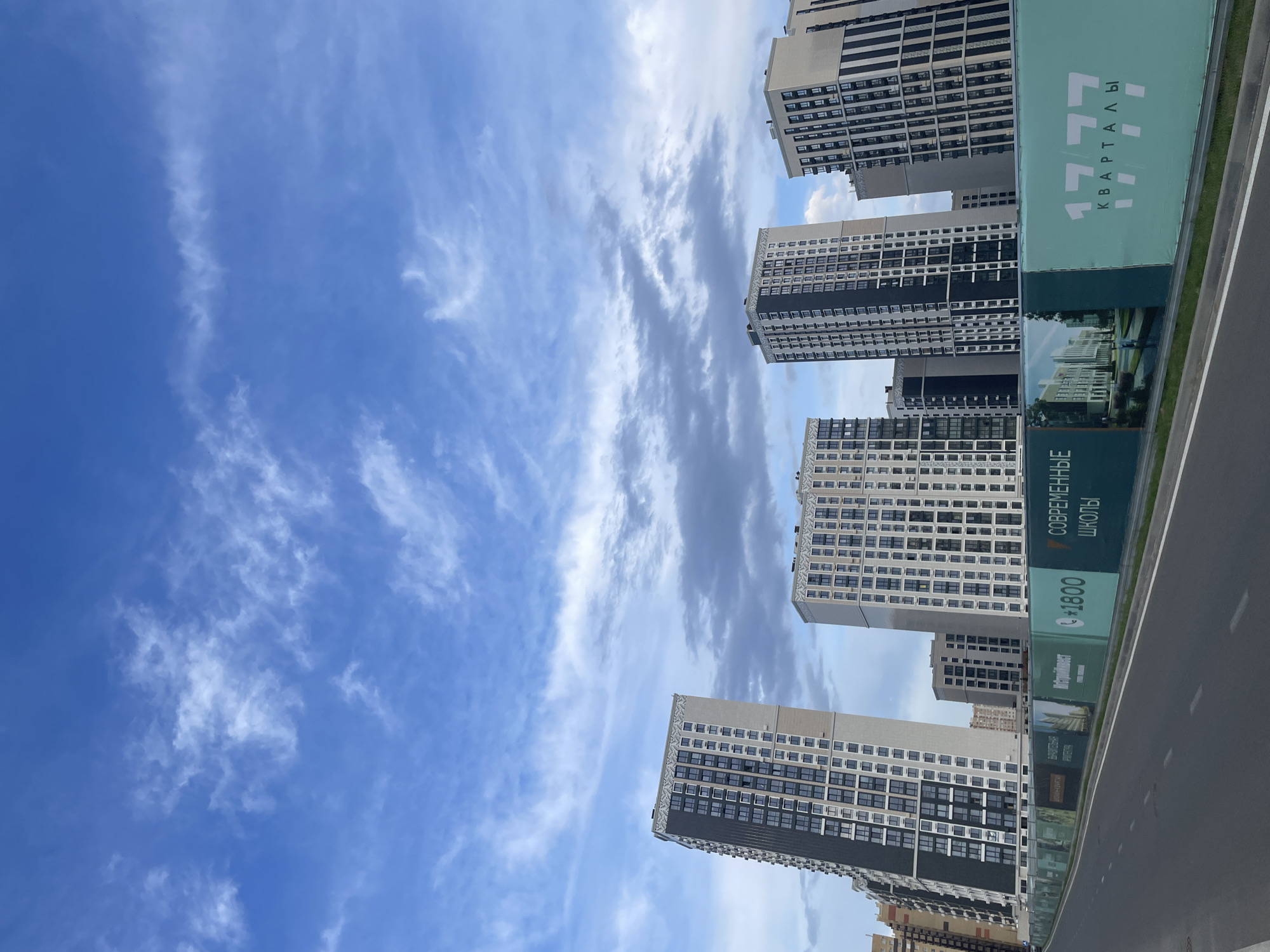 The image depicts a modern urban landscape with multiple high-rise apartment buildings under a partly cloudy blue sky. In the foreground, a grey asphalt road stretches into the distance, bordered by a green fence adorned with large banners. One banner prominently features the text "1777 КВАРТАЛЫ" in large white numerals and letters, suggesting a residential development. Another banner displays the text "СОВРЕМЕННЫЕ ШКОЛЫ" (Modern Schools) and a phone number "+1800". The architecture of the buildings is contemporary, characterized by repetitive window patterns and varied facade designs, with some exhibiting dark and light contrasting elements. The scene is devoid of any people or vehicles, creating a sense of stillness. The overall impression is one of a developing urban area, possibly on the outskirts of Stavropol, Russia, as indicated by the presence of new construction and promotional materials.