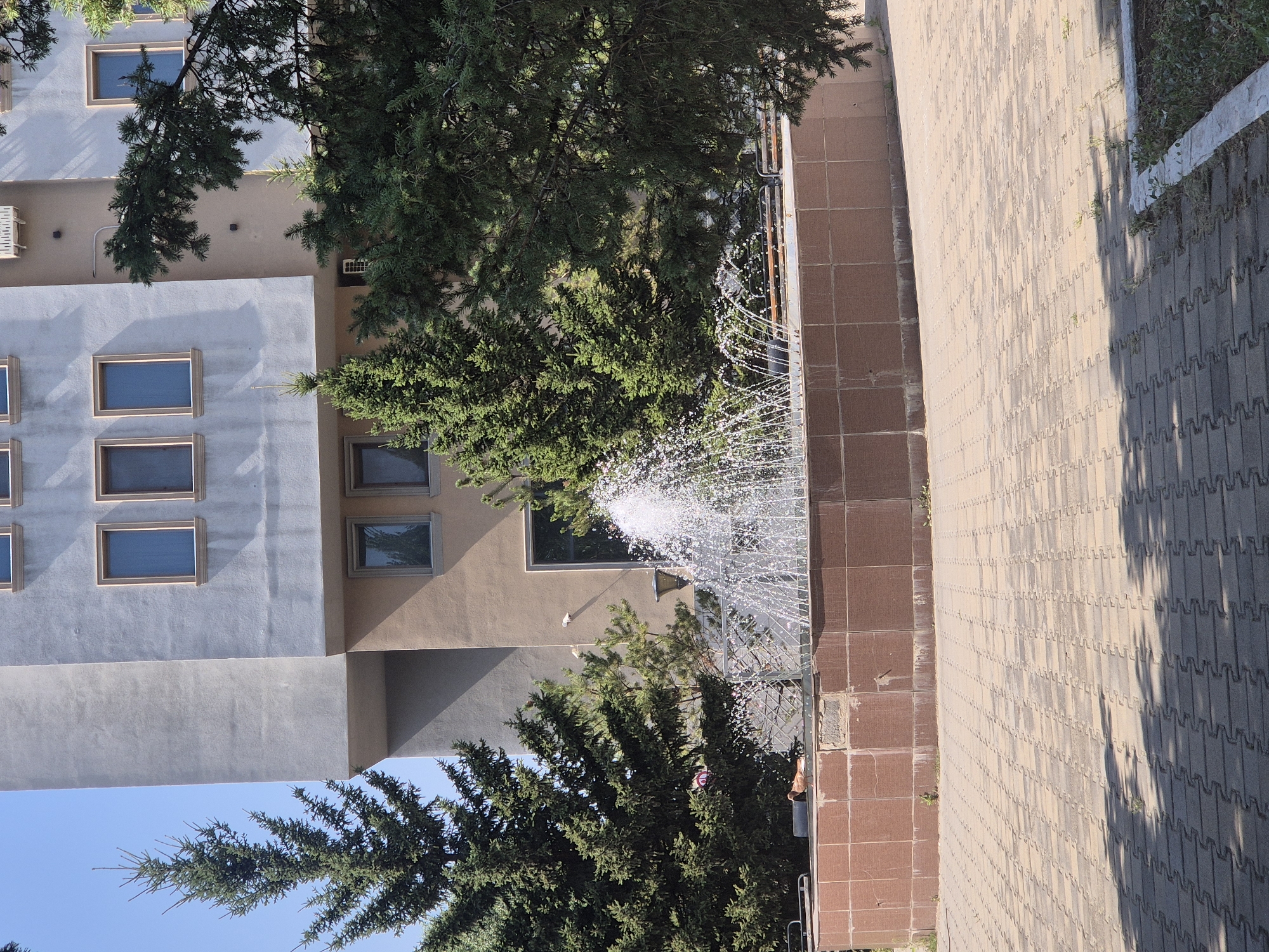 The image captures a daytime scene in Kokshetau, Kazakhstan, focusing on an outdoor public space with a fountain and a building. The foreground features a paved walkway with a repeating geometric pattern, casting shadows from an unseen source. To the left of the walkway, a circular fountain with a tiled base actively sprays water, creating a mist. Behind the fountain is a low wall, also tiled, with a single brown bird resting on its edge.
Adjacent to the fountain and walkway is a multi-story building, distinguished by its modern architectural style. The building has a light-colored facade with several rectangular windows, some framed in a light brown color. A portion of the building is painted a light gray, while another section is a muted beige. Some sections of the building have visible air conditioning units. Evergreen trees, some quite large, surround the area, with their branches extending into the frame, partially obscuring the building. The sky is clear and blue. The overall impression is that of a well-maintained urban park or plaza area adjacent to a contemporary building.