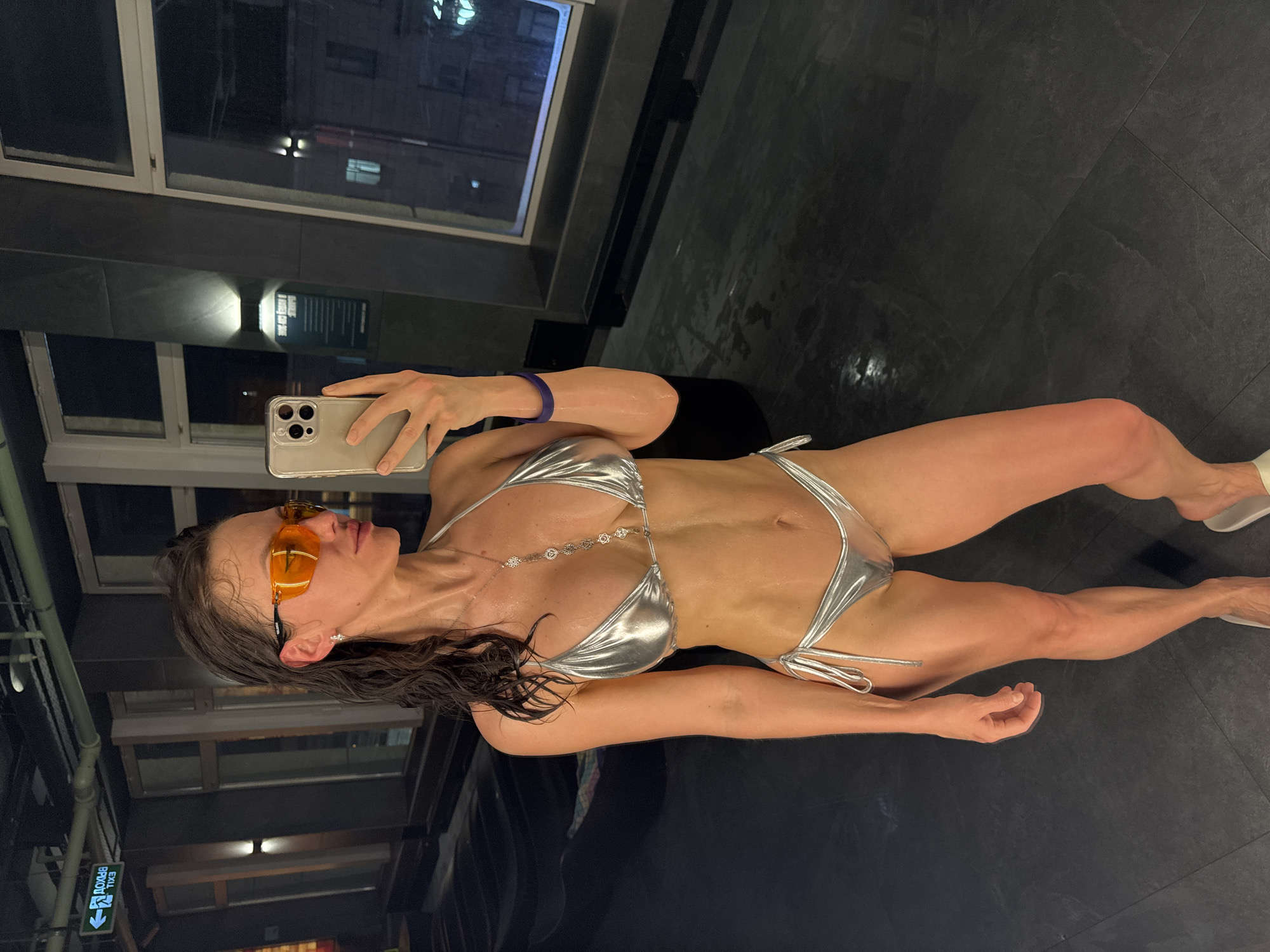 A full-body mirror selfie captures a woman in a sleek, dimly lit indoor setting, likely a spa, pool area, or changing room, characterized by dark, reflective tiled surfaces. The image is oriented vertically, showing the woman standing.
The woman, with wet, dark hair, wears an iridescent silver metallic bikini. She has a delicate necklace with circular pendants around her neck and a purple wristband on her left arm. Her eyes are obscured by large, orange-tinted sunglasses. She is holding a light-colored iPhone with a clear case in her right hand, angled to take the photo. Her physique appears toned. White sandals or slides are visible on her feet.
The background features dark, reflective walls and a floor with visible tile lines. A large window occupies the upper portion of the frame, showing a dark exterior with faint lights, suggesting it is nighttime. The window reflects elements of the room, including a partially visible "EXIT" sign with an arrow pointing left, located in the lower-left corner of the reflection. Another faint, inverted reflection of text, possibly a sign, is seen on a wall panel but is indistinct. The overall ambiance is modern and somewhat upscale.