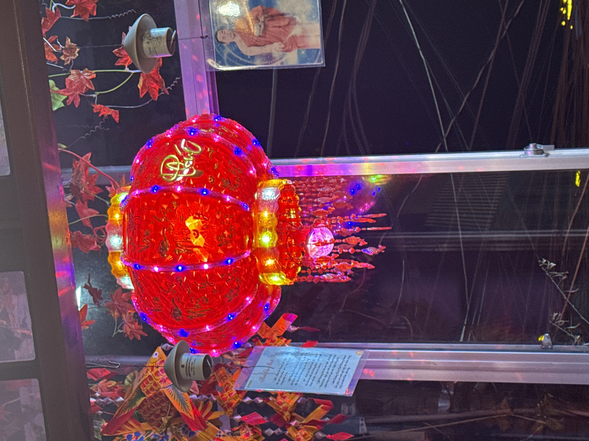 This nighttime image, captured in Da Lat, Vietnam, prominently features a large, illuminated red lantern at its center, adorned with gold script and embellished with blue, yellow, and green LED lights, along with dangling decorative elements including small, iridescent fish. To the left of the main lantern, several colorful paper fish or carp decorations in red, yellow, and blue hues are visible, hanging from above. A small, light-colored, bell-shaped ceramic-like object is suspended near these paper fish. Further to the right and slightly behind the main lantern, a similar bell-shaped object is partially visible against a dark background adorned with scattered red leaf-like embellishments. A vertical metal support pillar runs through the lower-middle portion of the frame. To the far left, a translucent rectangular sign with written text is hanging. On the right side of the image, a framed picture displays a man in red monastic robes, presumed to be a Buddhist monk, holding a bowl, set against an ethereal, cloudy background. The scene is illuminated by the lantern's glow and other ambient artificial lights, indicating an evening setting. The overall environment suggests a decorative outdoor or semi-outdoor space, possibly a shop entrance or temple area.