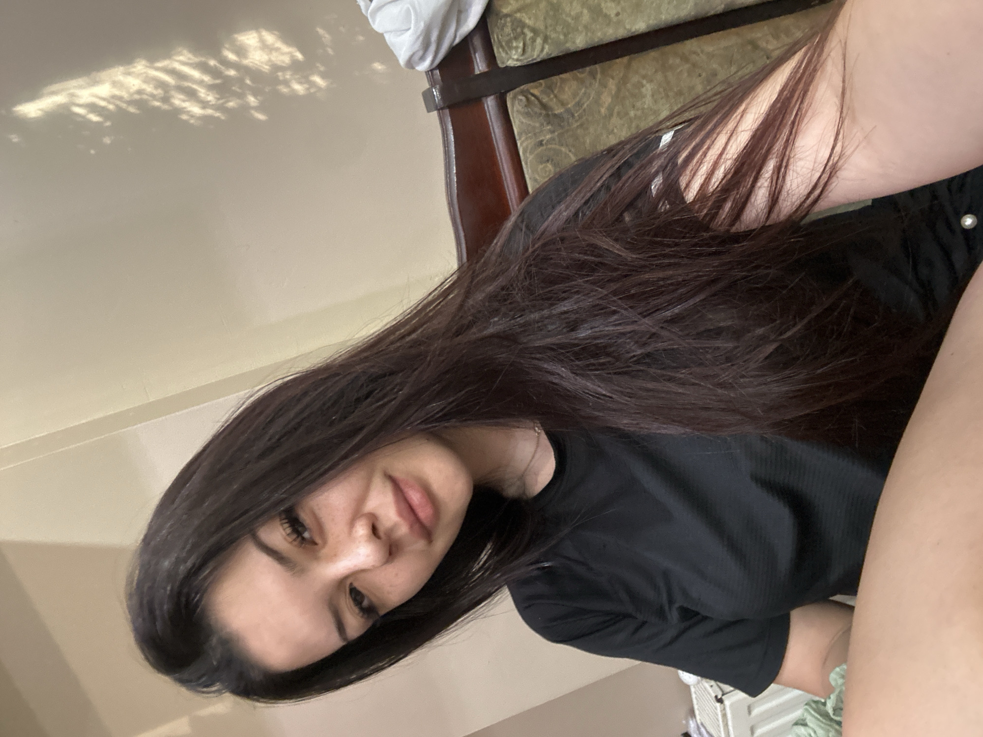 A young woman with long, dark hair and freckles is captured in a close-up selfie. She is wearing a black t-shirt and has makeup on her eyes and lips. The background appears to be an indoor setting, possibly a home, with a textured wall and a glimpse of a wooden chair with upholstery. There is also a radiator visible in the lower left corner. The lighting suggests natural light filtering in, casting shadows on the wall. The image does not provide any specific information about the city of Sliven, Bulgaria.