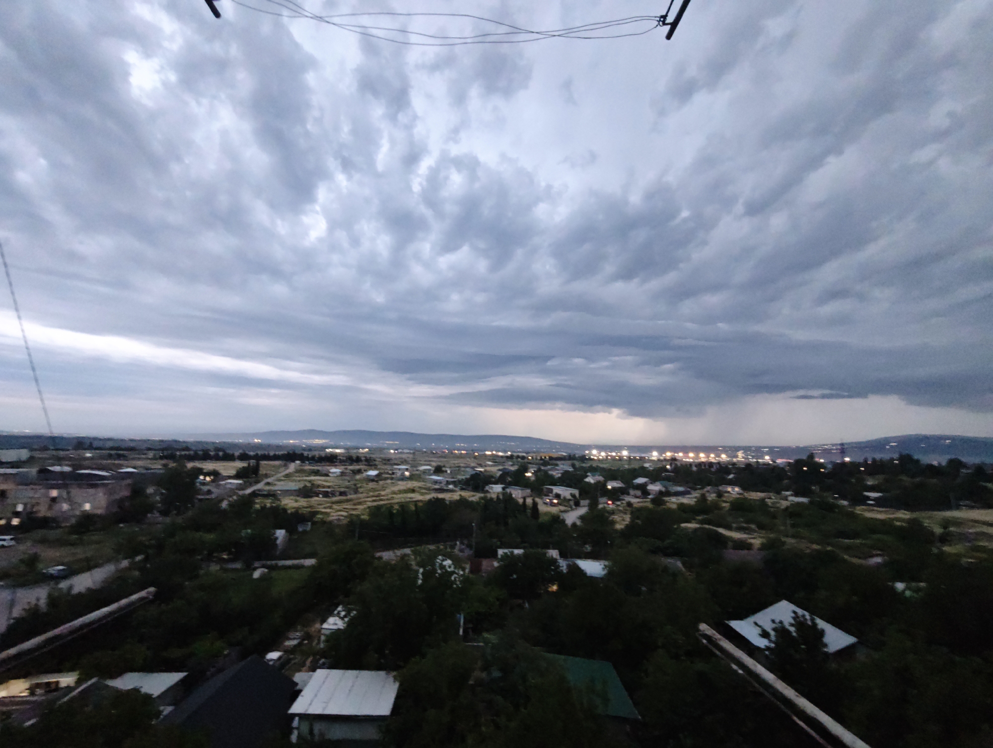 This is an aerial, wide-angle photograph taken at dusk, overlooking a suburban area of Tbilisi, Georgia. The sky is filled with heavy, dark grey clouds, suggesting an impending storm or recent rain, with a visible rain shower in the distance over the cityscape. Below the dramatic sky, a sprawling landscape unfolds, characterized by a mix of residential buildings, scattered trees, and undeveloped, grassy hills.
In the foreground, several houses with dark roofs are visible, some appearing aged with corrugated metal or weathered shingles. Further into the mid-ground, the residential architecture becomes more varied, with some buildings exhibiting more modern designs, and a few industrial-looking structures. Numerous trees are interspersed throughout the scene, some appearing dense and mature, contributing to a somewhat overgrown feel in parts.
The background reveals the distant lights of the city of Tbilisi, twinkling against the darkening horizon, suggesting a densely populated area. The terrain appears to be hilly, typical of Tbilisi's topography. The overall impression is of a somewhat unkempt but lived-in urban fringe, under a moody, atmospheric sky. There are no people or vehicles clearly discernible in the immediate foreground or mid-ground. The image captures a specific moment in time, likely during twilight hours.