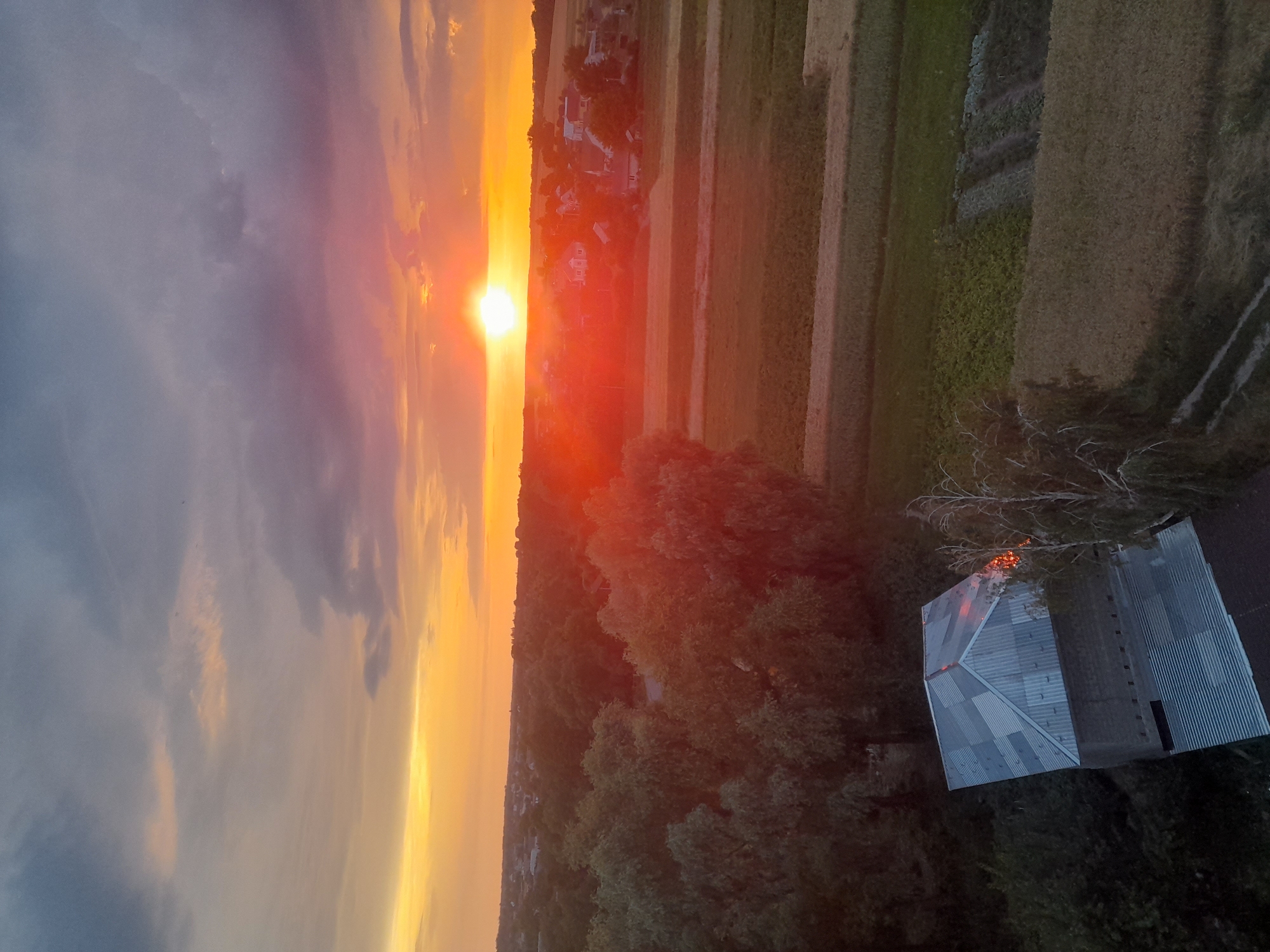 An aerial drone shot captures a sunset over the outskirts of Borodianka, Ukraine. The sky is a dramatic canvas of oranges, yellows, and purples, with thick clouds streaking across the horizon. The sun, a brilliant orb, is low in the sky, casting a warm glow over the landscape. In the foreground, a dilapidated, corrugated metal-roofed structure is visible, with some evidence of damage or disrepair. The surrounding area is a mix of trees and agricultural fields, with a small village nestled in the middle distance. The overall impression is one of a rural setting bathed in the warm, fading light of dusk, with a sense of quiet and possibly abandonment.