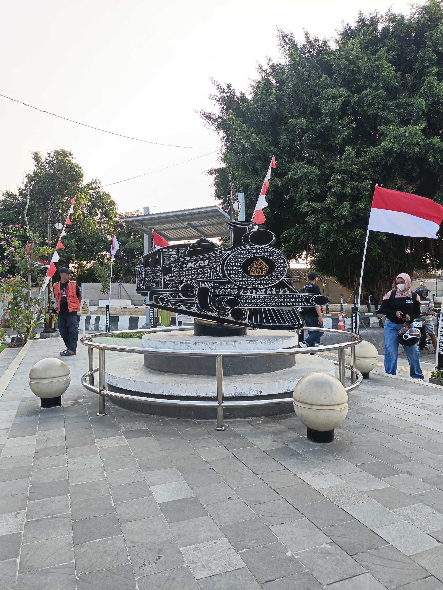 The image shows the iconic locomotive monument in the city of Klaten, Indonesia. The monument, which is a replica of a steam locomotive, is located in a public square with a paved walkway. A large tree with lush green foliage stands behind the monument, and Indonesian flags are displayed on poles around the square. Several people are visible in the image, including a man in an orange vest standing near the monument, a woman in a hijab and jeans, and another person on a motorcycle. The sky is clear and the lighting suggests it is late afternoon. The overall scene depicts a well-maintained public space with a historical monument that is a point of attraction for locals and visitors.