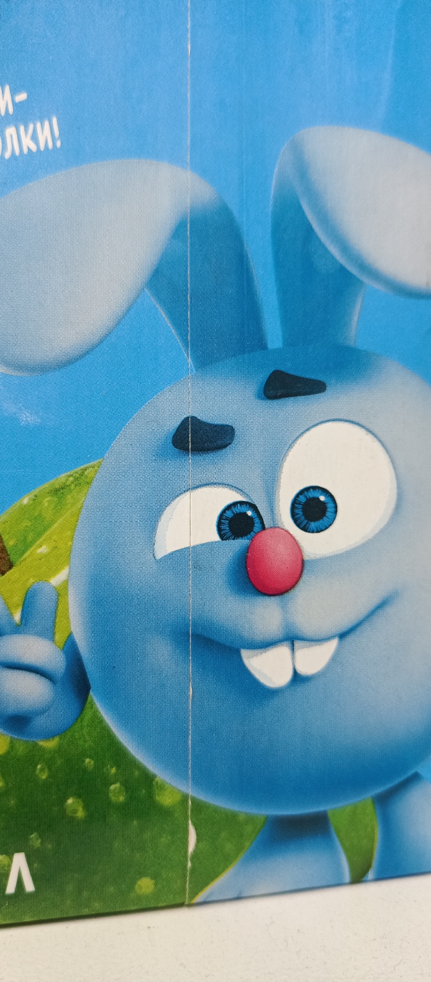 The image is a close-up shot of a blue cartoon rabbit's face. The rabbit has large, bright blue eyes with black pupils, a pink nose, and two prominent front teeth. Its ears are large and rounded, and one is partially visible at the top of the frame. The rabbit is depicted against a bright blue background, with a hint of green foliage and some water droplets in the lower left corner. Text in a white, sans-serif font is visible in the upper left, with the letters "М-" and "ОЛКИ!" discernable. A vertical crease runs down the center of the image, suggesting it's part of a larger printed material.