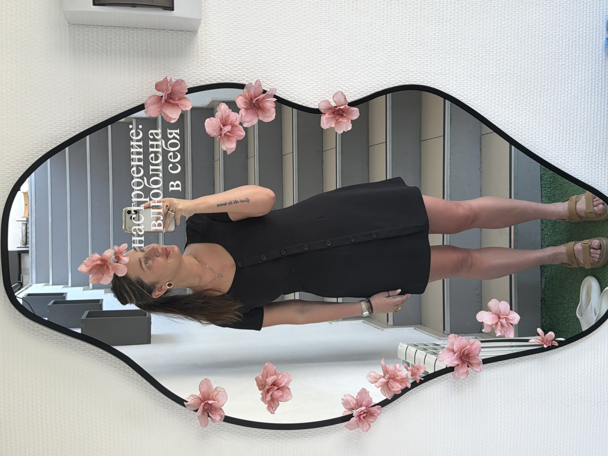 A young woman in a short black dress and tan sandals is taking a selfie in a decorative mirror. The mirror has a black frame and is adorned with pink fabric flowers. The text "настроение: влюблена в себя" (mood: in love with myself) is visible in white letters in the mirror's reflection.
The reflection shows stairs, grey boxes, and a patch of green turf. The location seems to be a stylish or decorative space, possibly a shop or an interior design studio. The specific location is in the city of Chaikovsky, Russia.