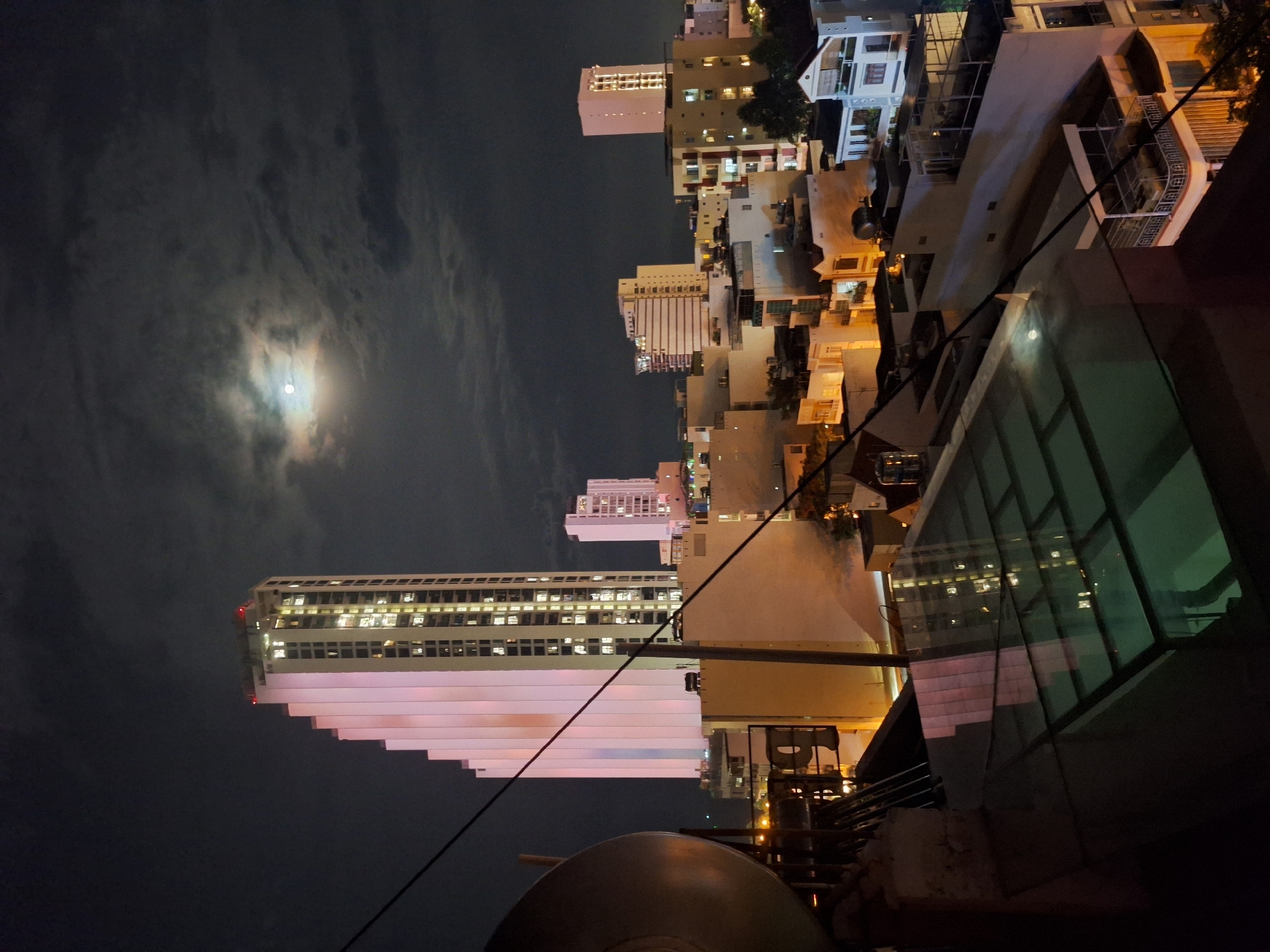 The image is a night time long shot of a city skyline in Nha Trang, Vietnam. The sky is dark and cloudy with a bright full moon peeking through. The main subject is a tall, modern skyscraper on the left, illuminated by pinkish-white lights. Its stepped design is clearly visible. Several other buildings of varying heights and architectural styles are spread across the cityscape, many of them showing illuminated windows, indicating occupied residential or commercial spaces. The foreground shows a dark rooftop with a reflective glass structure, and some electrical wires are visible, obscuring parts of the view. The overall impression is a bustling urban environment at night, bathed in moonlight.