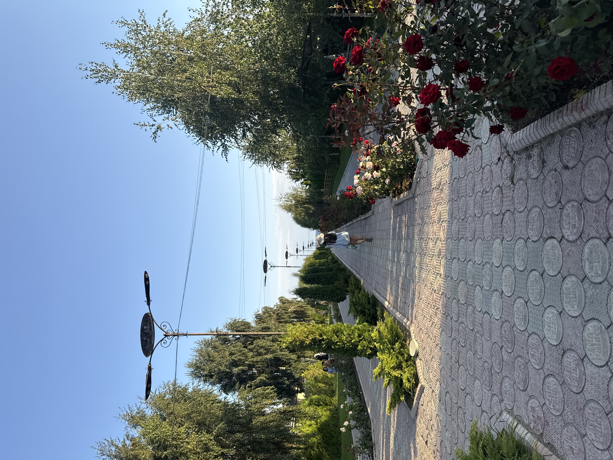The image depicts a paved pathway lined with rose bushes and manicured trees, likely in the city of Sary-Oy, Kyrgyzstan. A lone individual, seen from behind, walks away from the camera down the path, carrying a light-colored bag and wearing a light-colored hat. To the left of the path, several other individuals, including children, are visible in the distance near some trees and bushes. Streetlights are spaced along the pathway. The sky is clear and blue, indicating fair weather. The pathway itself is constructed with decorative patterned paving stones, some of which feature circular inlays with what appears to be text, possibly identifying the location as "Asia Palace." The scene suggests a public park or garden area.