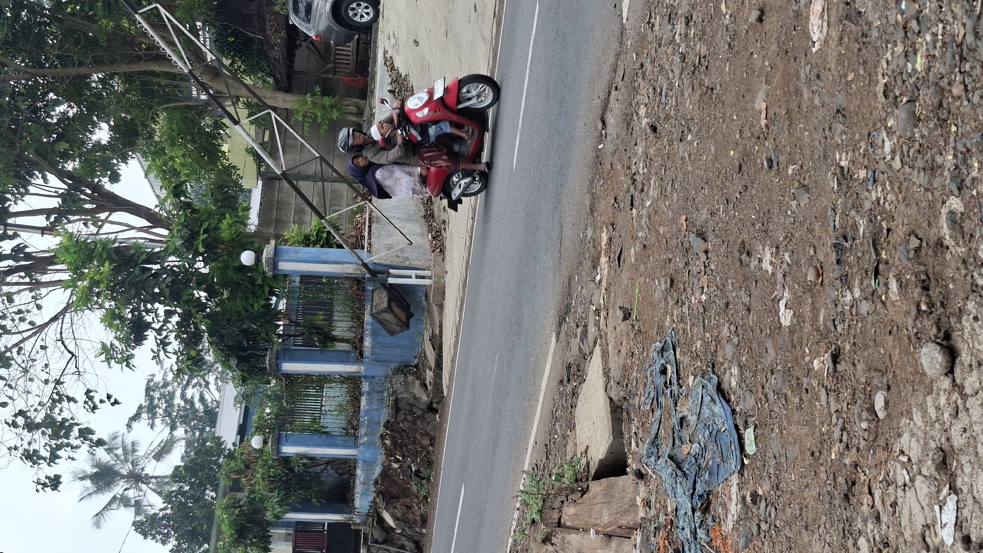 In Cilaku, Indonesia, a red scooter travels on a paved road, carrying three individuals. The driver, a woman wearing a helmet, is in the front. Behind her, a woman in a hijab is seated, with a young child in front of her. The scooter is positioned on the right side of the road, moving away from the camera.
To the left of the road, a blue wall with white trim runs along the property line. In front of the wall, there is a pile of debris consisting of rocks and soil. A bent metal pole, possibly part of an electrical pole or street sign, lies across the road, its upper portion angled upwards and to the right. The ground on the side of the road is uneven, with patches of dirt, rocks, and scattered debris, including a crumpled blue tarp.
In the background, there are trees, some with lush green foliage, and a palm tree on the left. Further back, a gray car is parked on the right side of the road, near a building with a thatched roof structure. The sky is overcast. The overall scene depicts a somewhat disheveled environment with the damaged pole suggesting a recent event or ongoing issue.